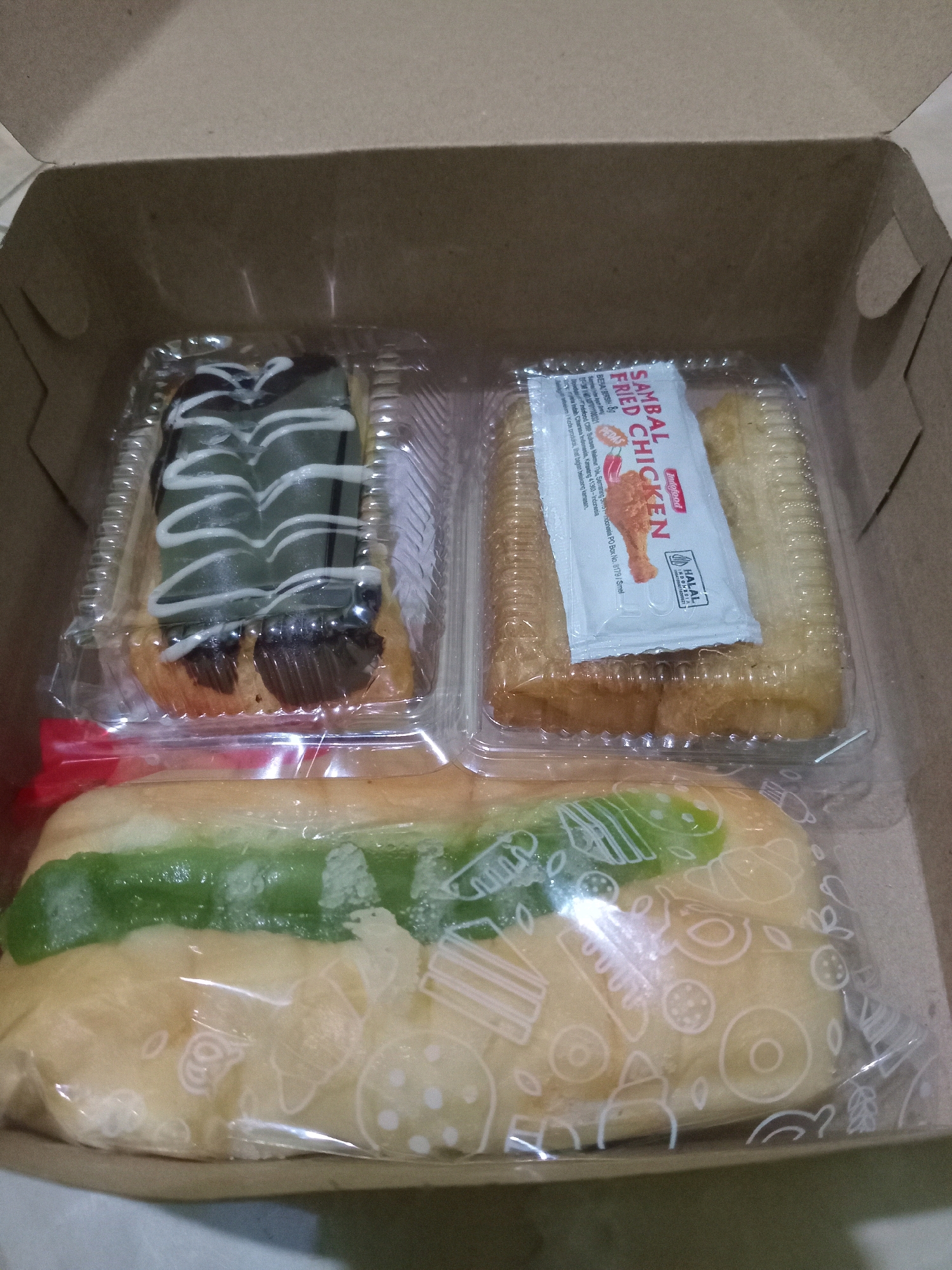 A brown cardboard box contains three food items, likely for takeout or delivery. In the top left is a pastry in a clear plastic container, topped with green frosting and white icing. To its right, another clear plastic container holds what appears to be fried breaded chicken pieces and a small packet of "Sambal Fried Chicken" sauce, also labeled with a "HALAL" certification. At the bottom of the box, a long pastry, possibly a bun, is wrapped in clear plastic and filled with a green substance, resembling a sweet filling. The overall setting suggests a casual food service environment, possibly in Randuagung, Indonesia, given the product labeling.