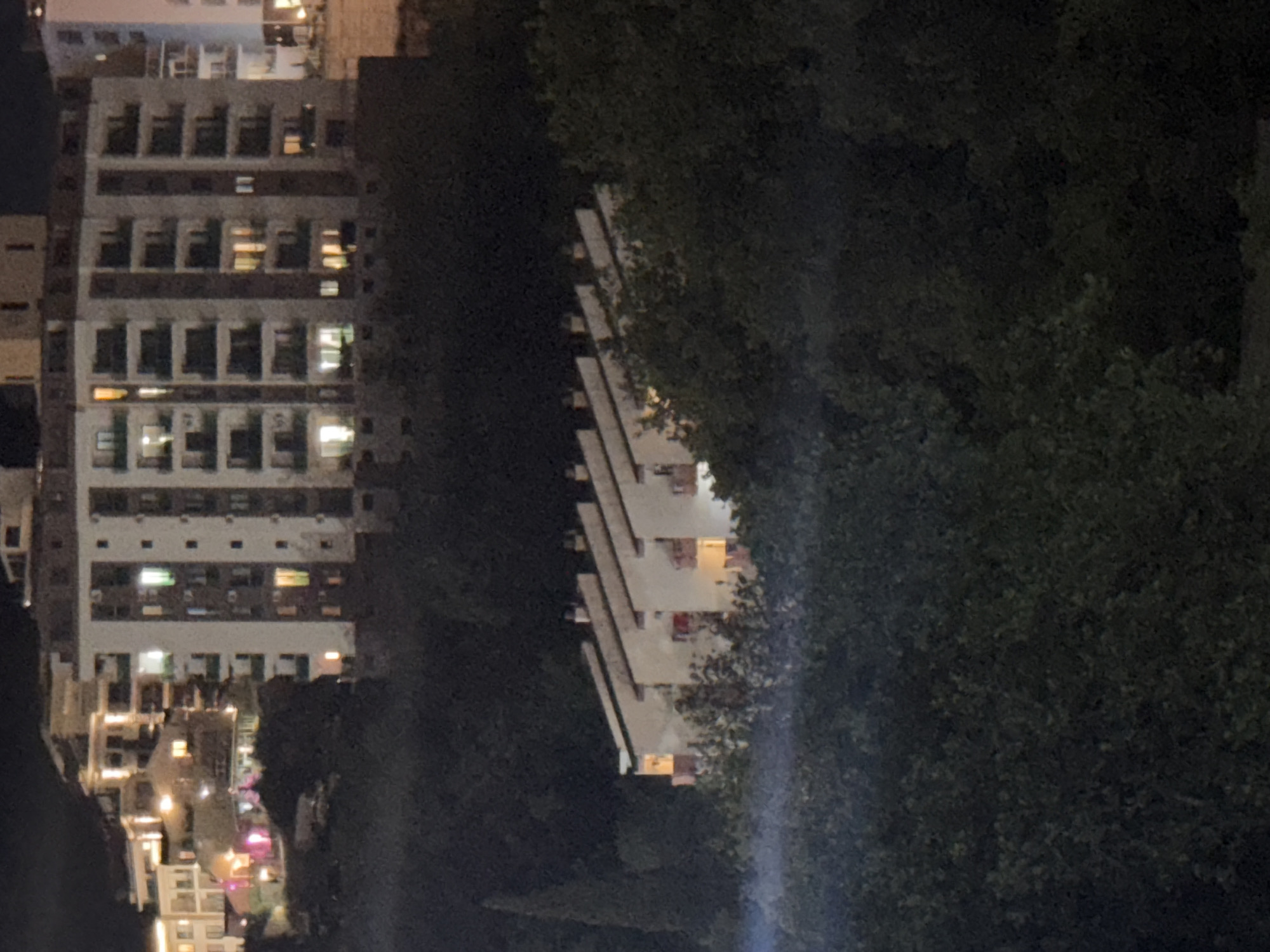 The image depicts an aerial view of a residential area at night in Boreti, Montenegro. The scene is dominated by buildings and dense vegetation.
In the foreground, a large expanse of dark green foliage, likely trees and bushes, occupies the lower portion of the frame.
Behind the foliage, a row of attached houses with slanted roofs is visible. These houses appear to be illuminated from within, with warm yellow light emanating from some windows and balconies. The houses have a light-colored facade and red accents on the balconies.
Further back and higher up, a larger, multi-story apartment building is prominent. This building is also illuminated with multiple lit windows, showcasing various shades of light including yellow, green, and white. The building has a modern appearance with balconies and vertical design elements. To the left of this apartment building, several other buildings of varying heights and architectural styles are visible, also showing signs of nighttime illumination. The overall impression is of a developed hillside community at dusk or night.