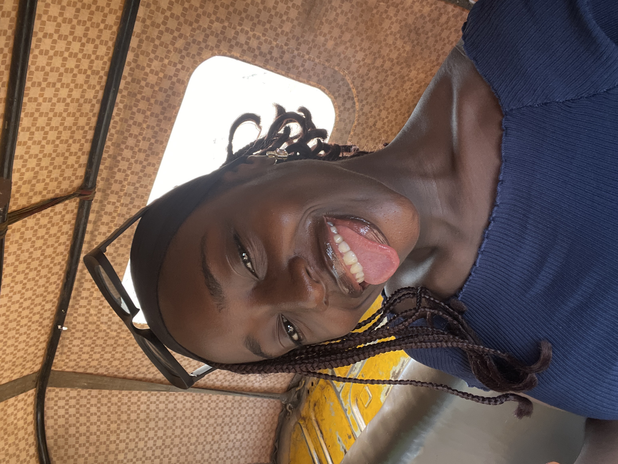 A young Black woman with dark skin and braided hair is inside a vehicle. She is wearing a dark blue off-the-shoulder top and has sunglasses perched on her head. She is sticking her tongue out at the camera and smiling. The interior of the vehicle has a textured, tan-colored ceiling and a yellow seat. The scene appears to be a candid photograph taken from a low angle, likely from within the vehicle. The background suggests an outdoor environment, but specific details of Abuja, Nigeria, are not identifiable from the image alone.
