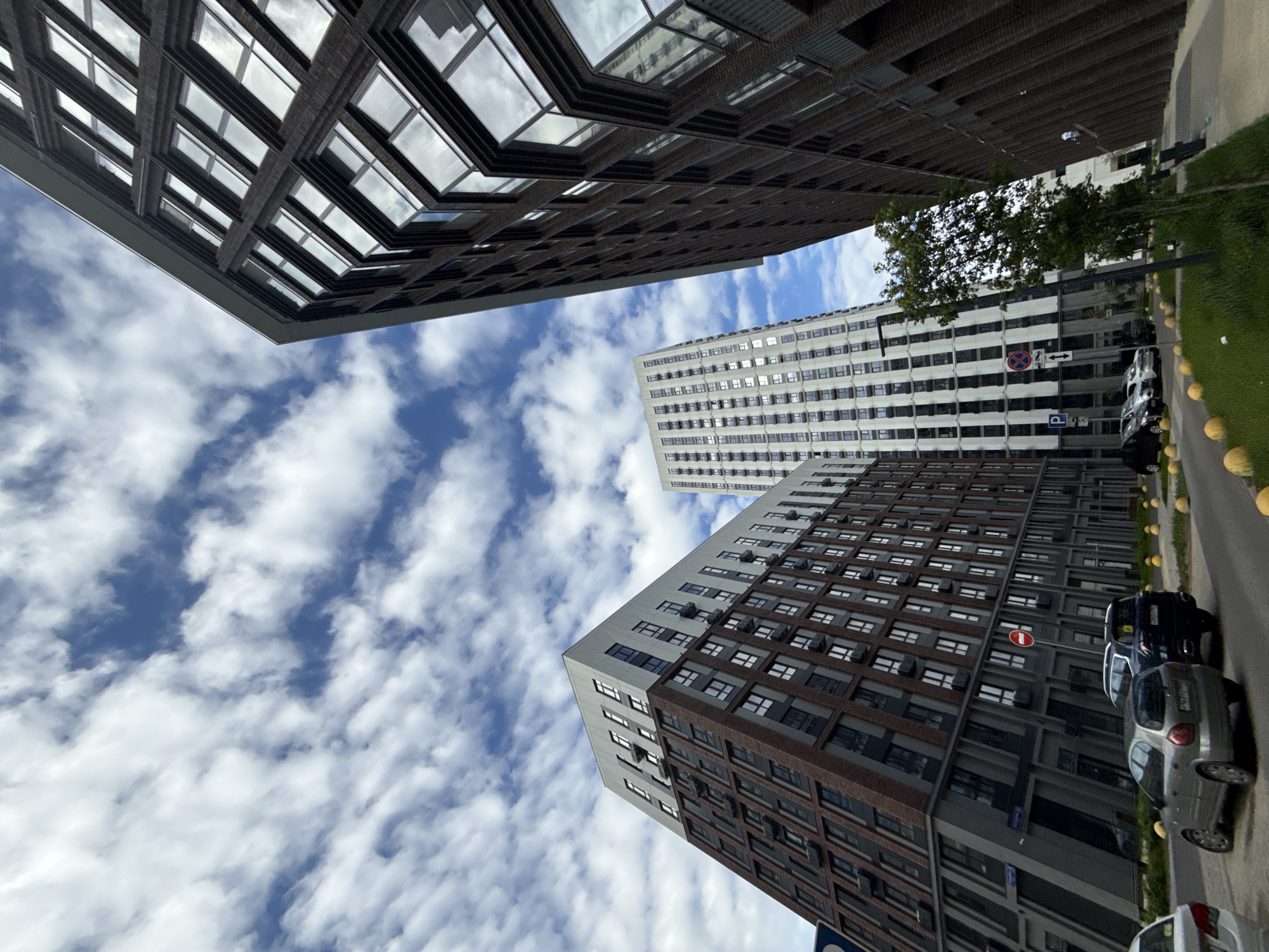 The image is taken looking up at tall buildings in what appears to be a city street. Three buildings are visible, varying in design. The building on the left is characterized by its dark brown exterior with a grid-like window pattern, giving it a modern architectural style. The building in the center is taller, with a predominantly white facade and numerous windows. The building on the right features a mixed facade of brown and white. The sky visible between the buildings is a bright blue with fluffy white clouds.
On the ground level, there is a street with cars parked along the side. Yellow spherical bollards line the street separating it from a grassy area. Street signs are visible on the buildings.
The location is identified as Moscow, Russia based on architectural style and street signs.