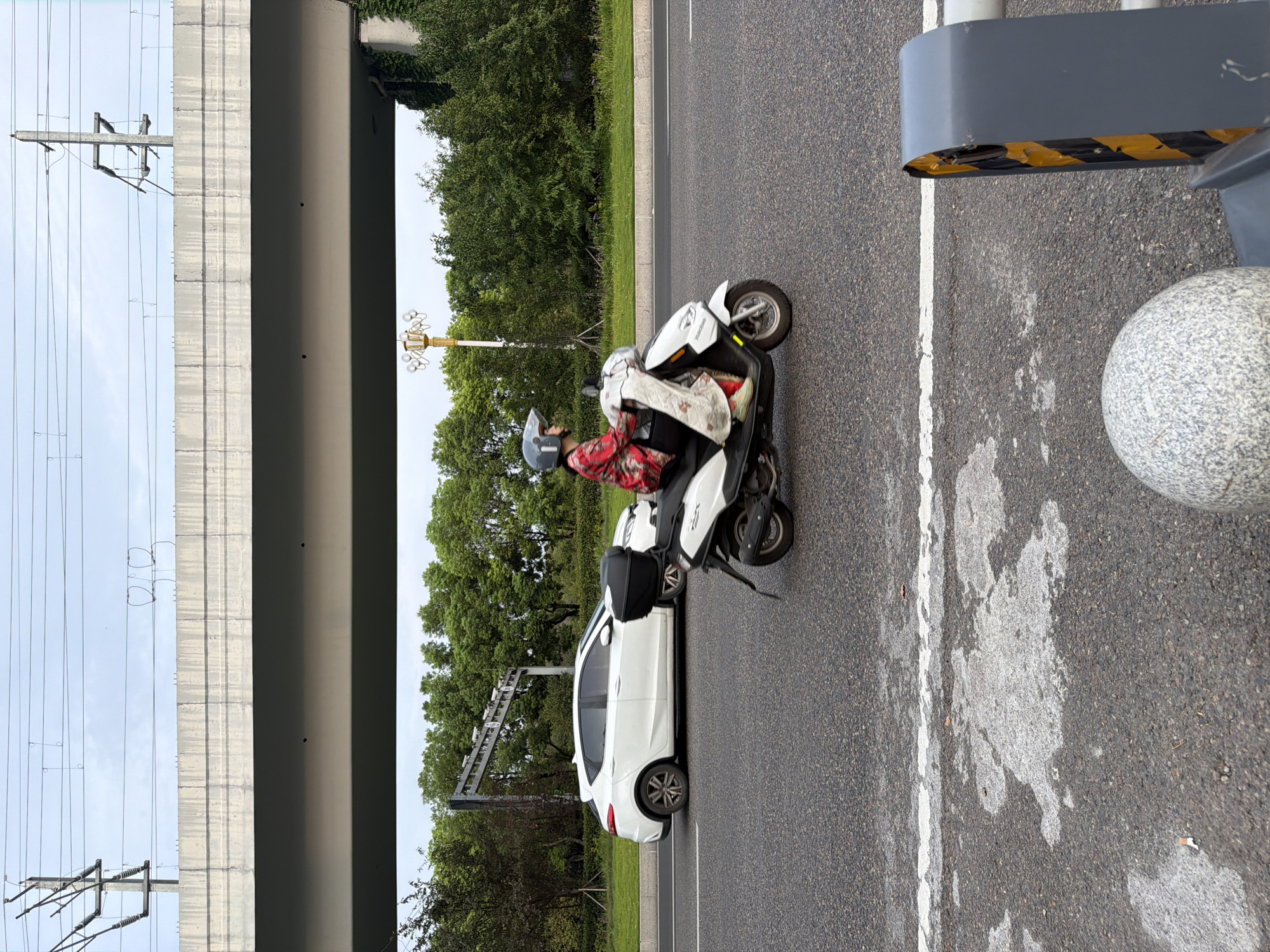 In Jiangjin, China, a person wearing a red and white patterned jacket and a helmet is riding a white scooter on a paved road. The scooter has a black storage box on the back. A white car is visible to the left of the scooter. Overhead, a concrete overpass with power lines runs across the frame. The environment includes green trees and vegetation lining the side of the road. The sky is cloudy. The scene appears to be a typical urban street.
