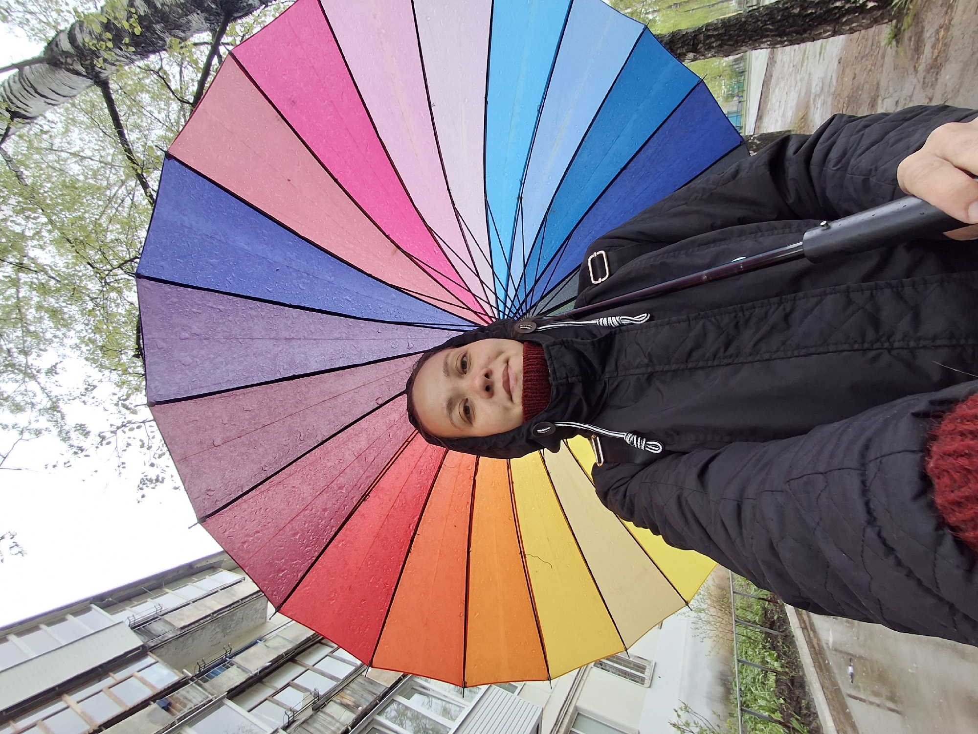 A person is holding a colorful umbrella in what appears to be a city environment, possibly Chaikovsky, Russia. The umbrella is rainbow-colored, with segments of red, orange, yellow, and other hues. Water droplets suggest it might be raining. The person is wearing a black jacket and a red sweater. The buildings and trees are visible in the background.