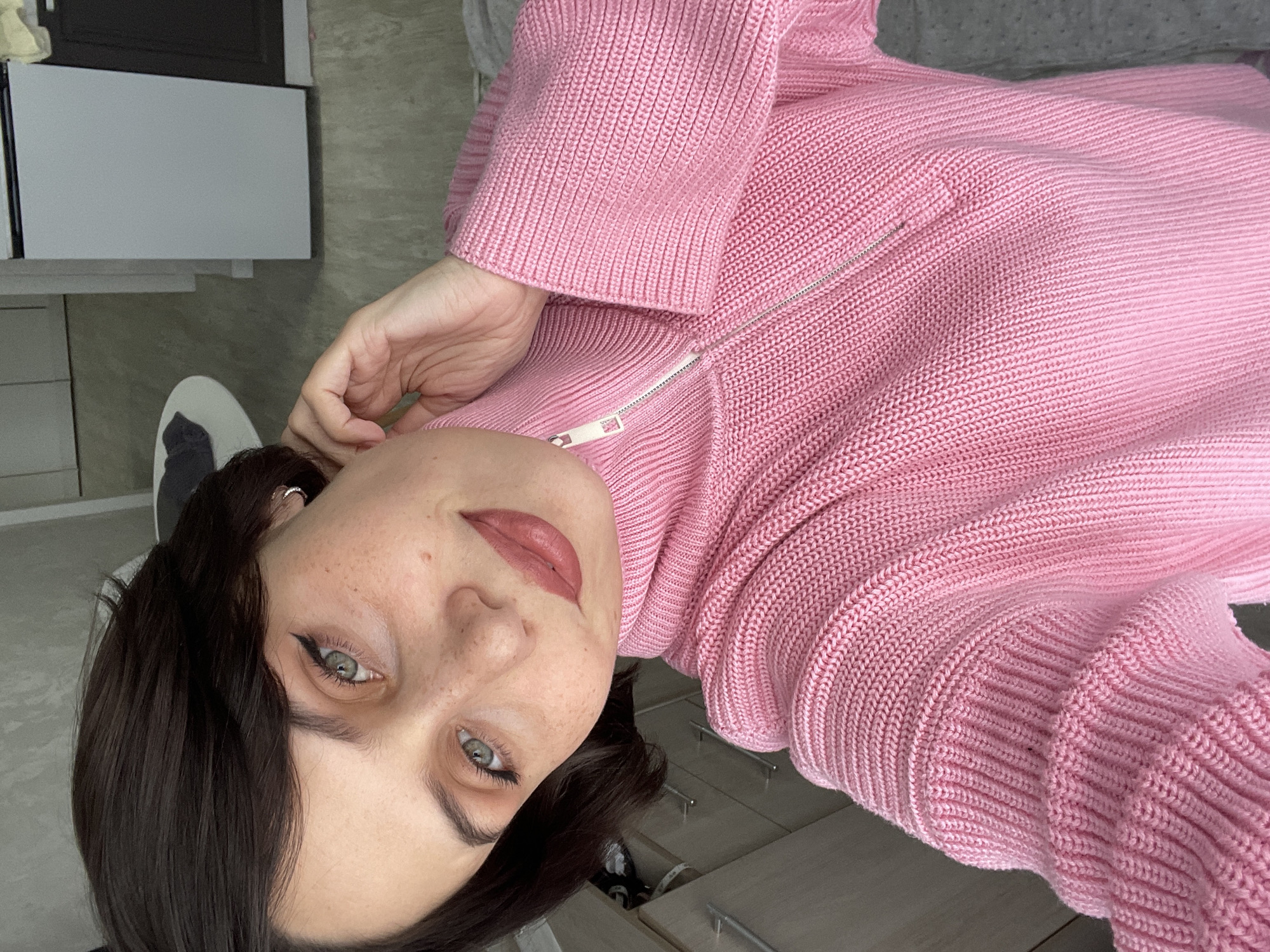 A woman with dark hair and light green eyes is wearing a pink sweater with a zipper. She has fair skin with freckles. The background shows a white cabinet, a partially visible white shoe with a dark tongue, and a gray wall. The image appears to be taken indoors. The location in the city of Izhevsk, Russia, is not apparent from the image.