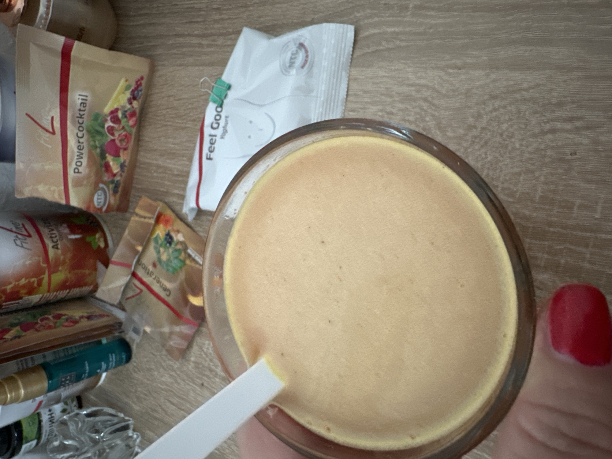 The image shows a close-up of a glass filled with an orange-colored smoothie. A white spoon is partially submerged in the smoothie. Behind the glass, several product packages are visible, including one labeled "PowerCocktail" and another that says "Feel Good Yoghurt." There is also a clear glass dish with a decorative pattern. The background appears to be a wooden surface. The image quality is moderate, with some blurriness. The scene is likely indoors, possibly on a countertop or table. There is no indication of the specific location within Krefeld, Germany.