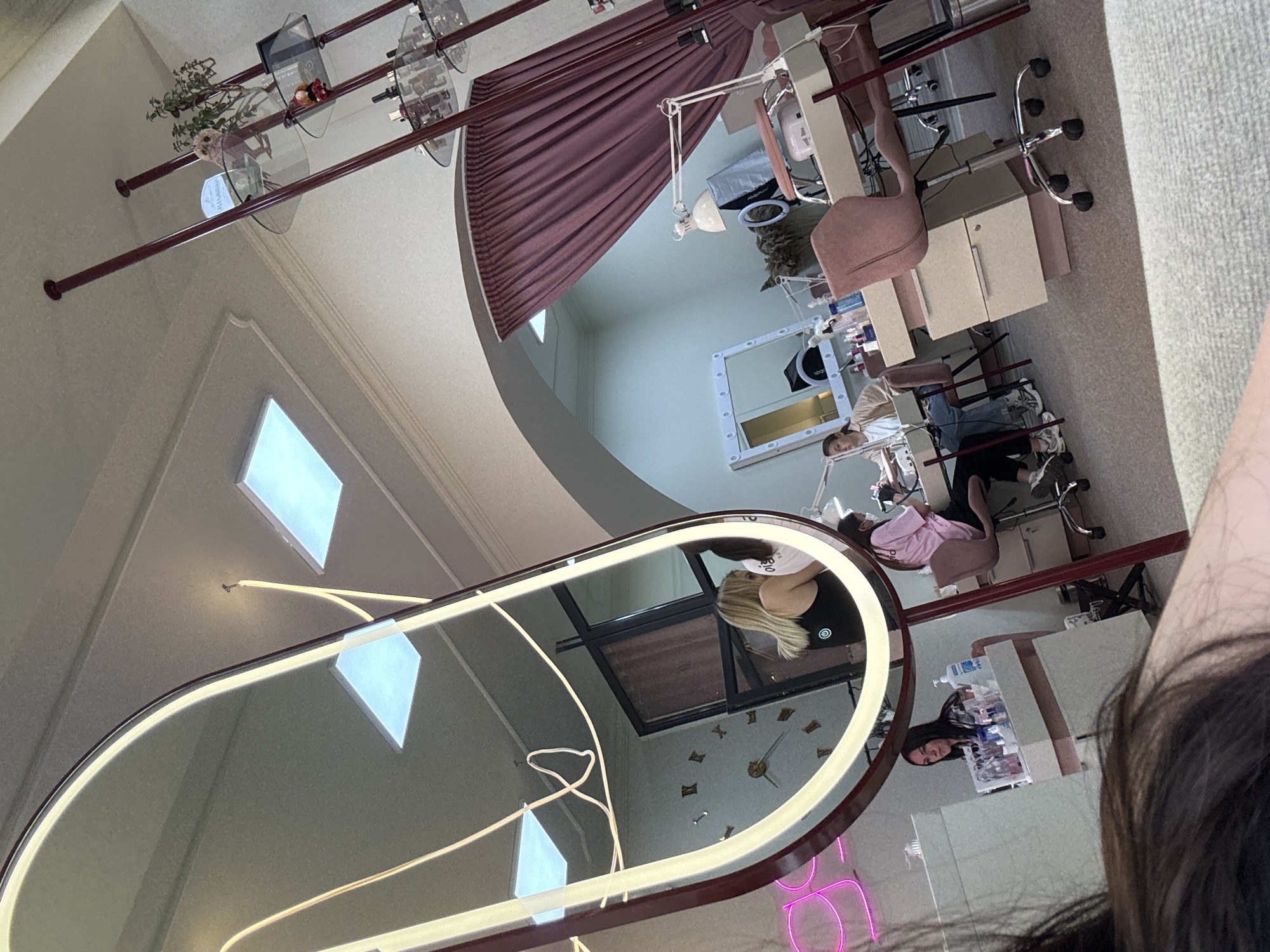 The image depicts the interior of a nail salon, likely in Chernivtsi, Ukraine, given the context. The scene is viewed from a low angle, looking across a carpeted floor towards several nail stations. Dominating the left side of the frame is a large, oval mirror with a bright, neon-like frame, reflecting portions of the salon, including a clock on the wall and people working at nail stations.
In the reflected image, one can observe a woman with blonde hair wearing a black tank top, and another woman with dark hair wearing a pink t-shirt. Several nail stations are visible, each equipped with a table, chairs, lamps, and various manicure tools and products. One station has a pink chair, and another appears to be occupied by a woman with dark hair in a pink shirt and jeans, seemingly a client. Further back, a woman in a light-colored jacket and jeans is seated at a nail station, her hands engaged in what appears to be a manicure service.
The salon environment appears modern and well-lit, with recessed ceiling lights. A tall, dark red shelving unit with glass shelves holding bottles and decorative items is positioned on the right side of the image. A pink neon sign with illegible text is partially visible on the far left. The overall impression is that of a busy, functional beauty salon.