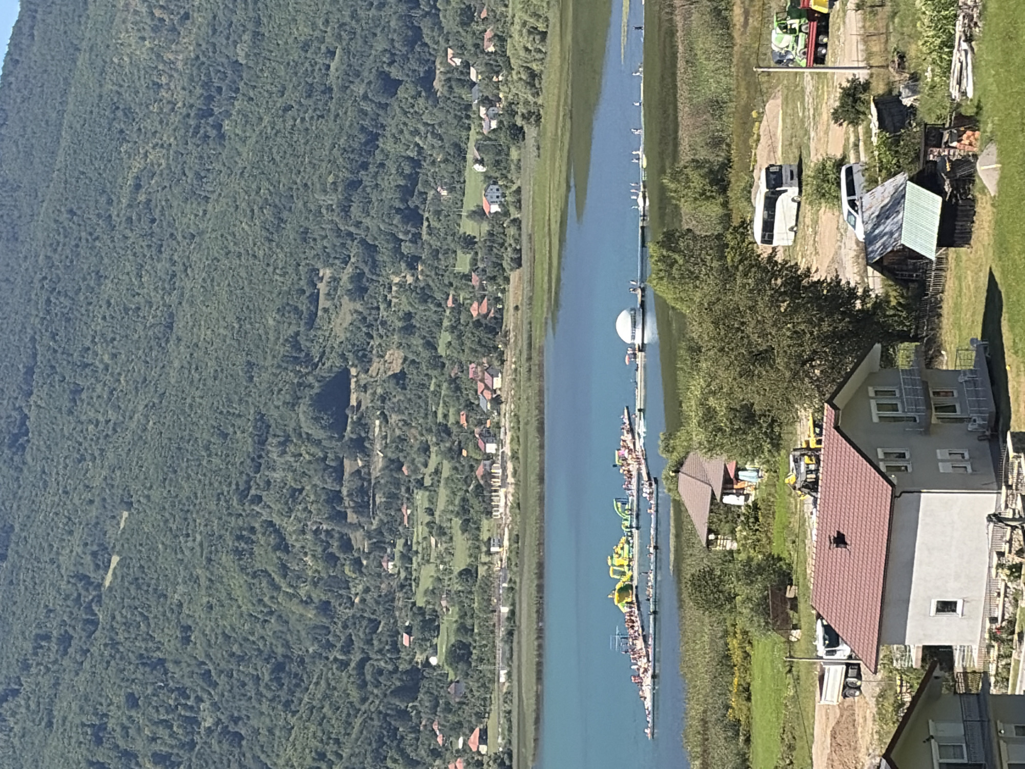 The image shows a landscape view of a lake with a green forested mountain in the background. In the middle ground, there is a body of bright blue water with a floating structure featuring inflatable water attractions and many people. Further back, there are a series of houses scattered on a green hillside. In the foreground, there are a few houses, some with red tiled roofs, and a road with a white bus and a white van. The scene appears to be in Plav, Montenegro, a location known for its natural beauty and lakes.