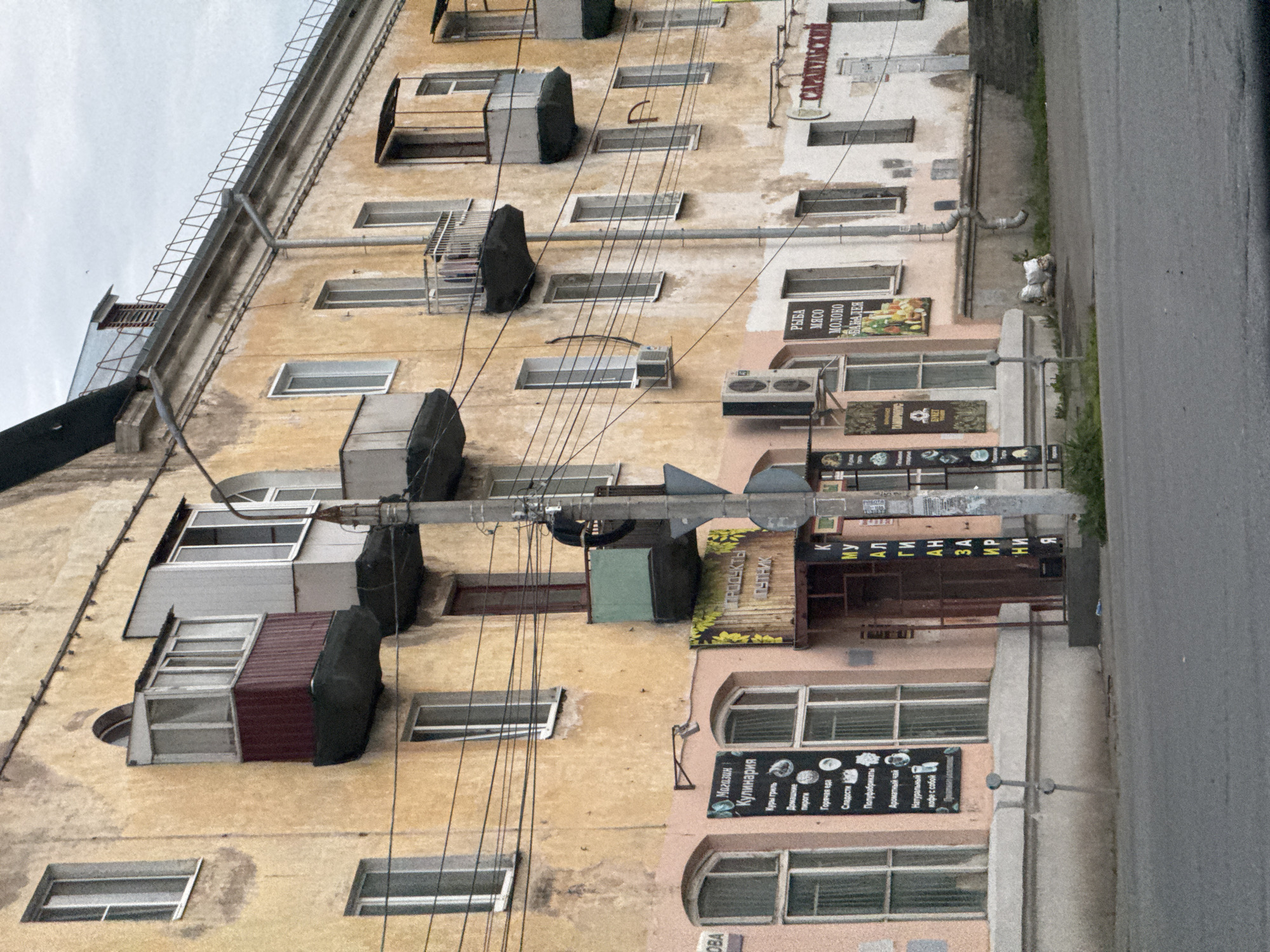 The image shows a weathered, yellow apartment building in Izhevsk, Russia, with multiple windows and balconies. The building appears to be aged, with peeling paint and some signs of wear. There are several shops on the ground floor, indicated by various signs. One sign, in Russian, says "Магазин Кулинария" (Shop Culinary), listing items like "Куры гриль" (Grilled chicken), "Домашние пироги" (Homemade pies), and "Сладости" (Sweets). Another sign shows "ПРОДУКТЫ ПУТНИК" (Traveler's Products). Further along, there's a sign for "РЫБА МЯСО МОЛОКО БАНАЛЕЯ" (Fish Meat Milk Banana). A store to the right has a sign that reads "САРАПУЛЬСКИЙ" (Sarapulsky). The street in front of the building is paved, and there is a sidewalk with some greenery. A utility pole stands prominently on the right side of the image. The sky is overcast.