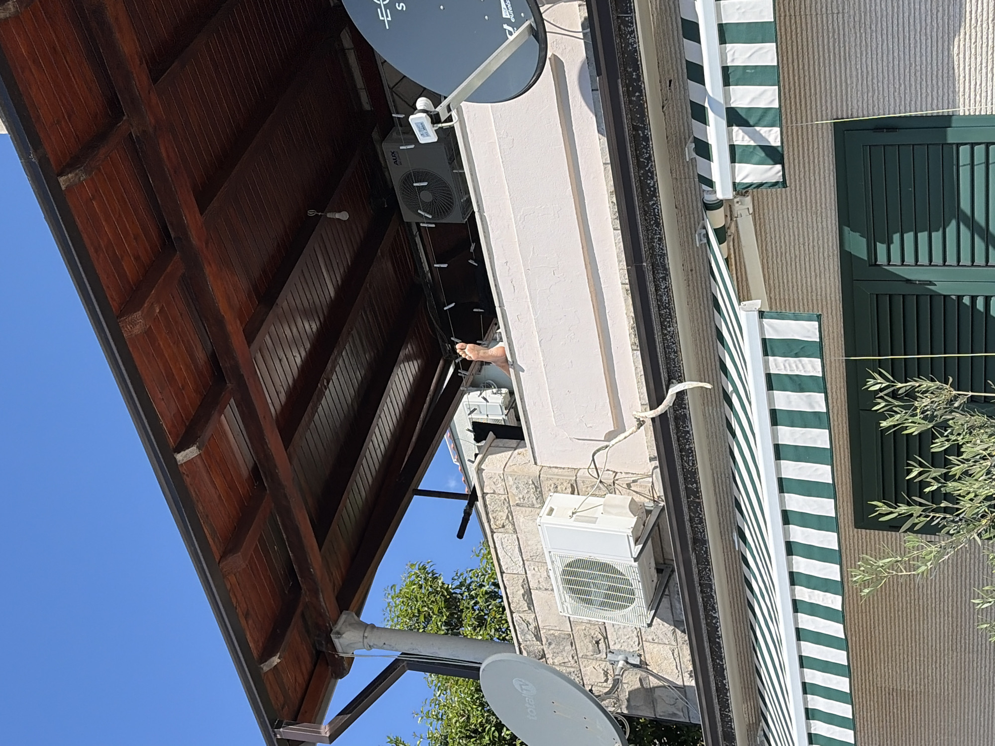 The image presents a partial view of a building exterior in Herceg Novi, Montenegro, under a clear blue sky. The structure features a wooden roof overhang with visible rafters. Two air conditioning units are mounted on the textured stone wall, with satellite dishes positioned above and below. A striped green and white awning partially covers a window with closed green shutters. A single foot extends from an unseen window. Vegetation is visible in the lower portion of the frame.