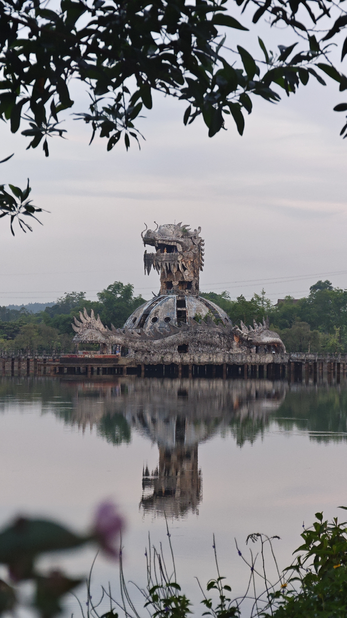 A full shot captures the Ho Thuy Tien Water Park, an abandoned water park structure designed to resemble a dragon, located in Thủy Xuân Ward, Vietnam. The dragon structure stands on a lake, its reflection visible in the water. The foreground features blurred foliage, while the background shows trees and a cloudy sky.