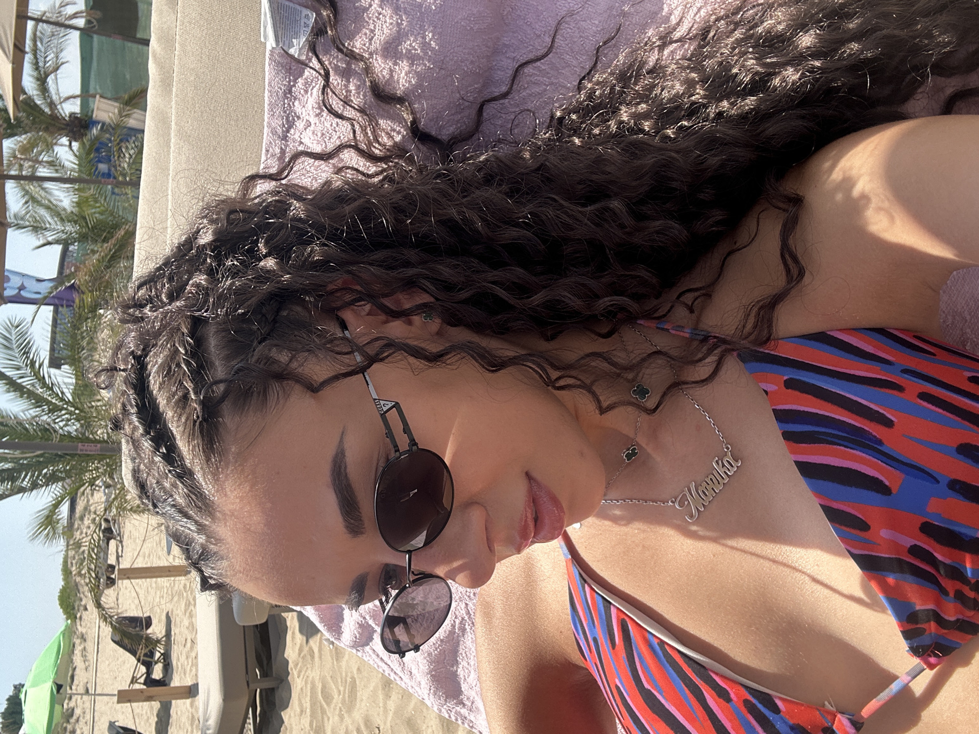 In the city of Nesebar, Bulgaria, a young woman with curly dark hair is lying on a beach chair. She is wearing sunglasses and a colorful bikini top. Her hair is styled in braids and is partially covering her face. A necklace with the name "Narmika" is visible around her neck. The background shows a beach with palm trees and umbrellas, indicating a sunny day. The woman appears relaxed and is enjoying the beach.