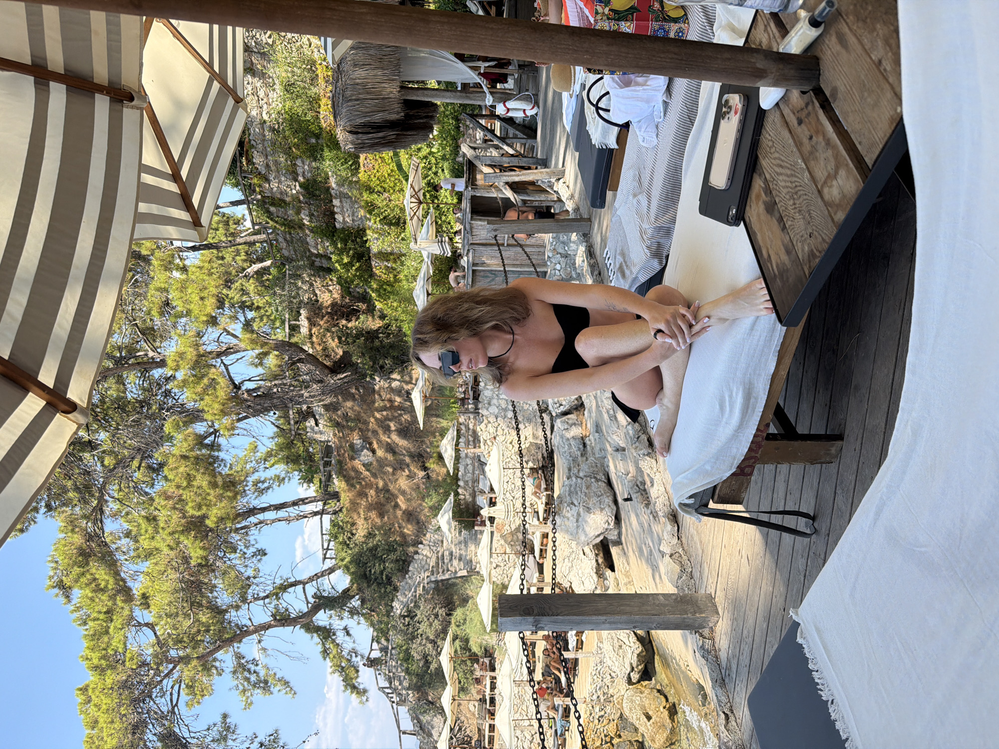 A woman in a black bikini sits on a beach chair, looking out at the water, on a sunny day in Fethiye, Turkey. She has sunglasses on and her legs are crossed. The beach is lined with lounge chairs and umbrellas. In the background, there are trees and a stone wall.