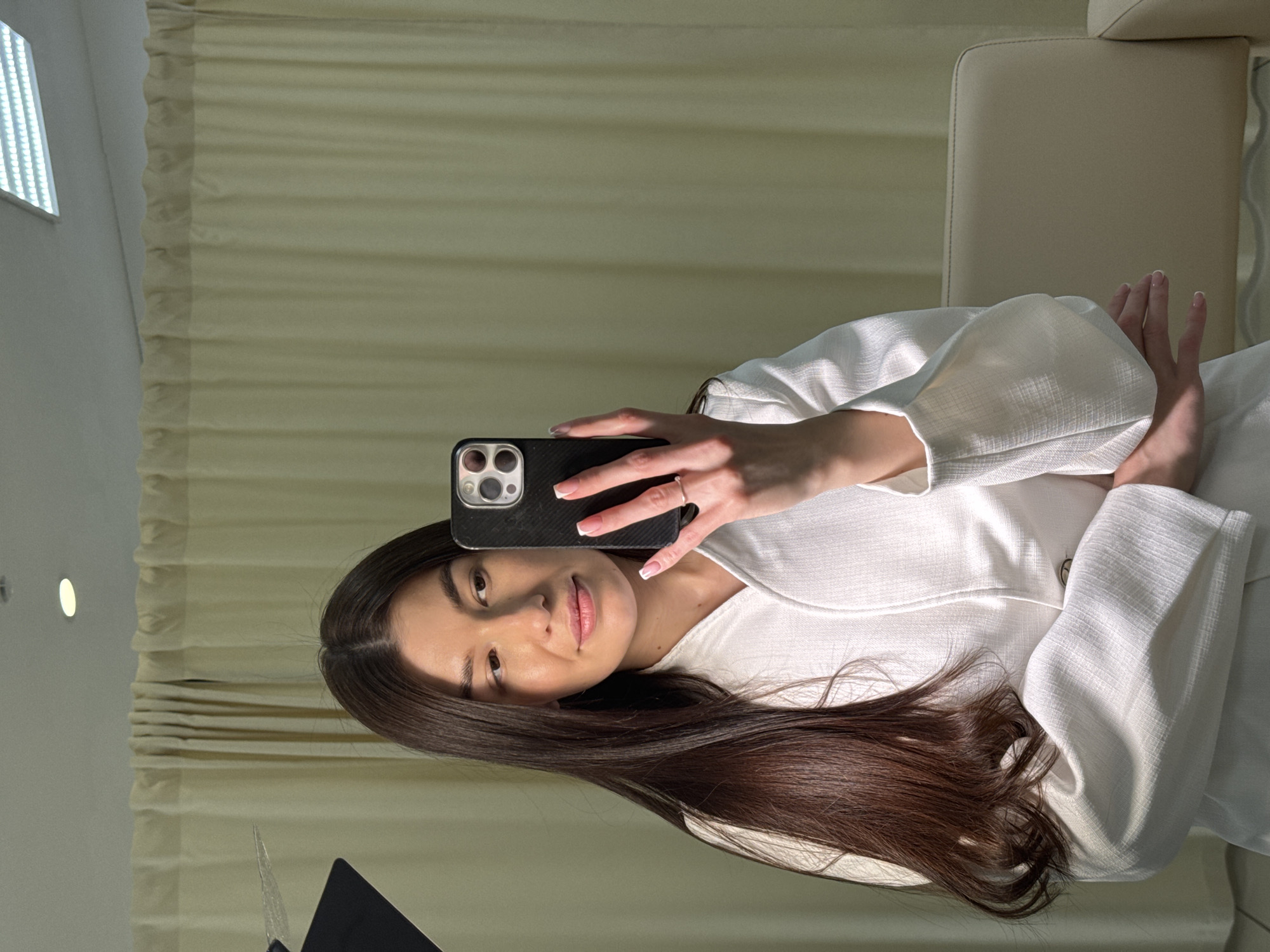 A young woman with long, dark brown hair and fair skin takes a selfie, holding a smartphone with a black, textured case in her right hand. She is looking directly at the camera with a subtle, composed smile. Her right hand displays a ring on her ring finger and nails with a French manicure. She is wearing a light-colored, possibly white or off-white, top or jacket with textured fabric and slightly puffed sleeves.
The setting appears to be indoors, potentially a home, hotel room, or studio, in Chaikovsky, Russia. The background features light beige or off-white curtains with horizontal folds, possibly Roman blinds, covering a window. To the upper left, a rectangular LED light panel is visible on a white wall or ceiling. To the right, part of a light-colored, possibly leather, armchair or headrest is present. The overall lighting is bright and even, with some natural-looking light illuminating her face. A dark, reflective surface, possibly a laptop screen or tablet, is partially visible in the bottom left corner. The image conveys a calm and bright atmosphere. No discernible text is visible in the scene.