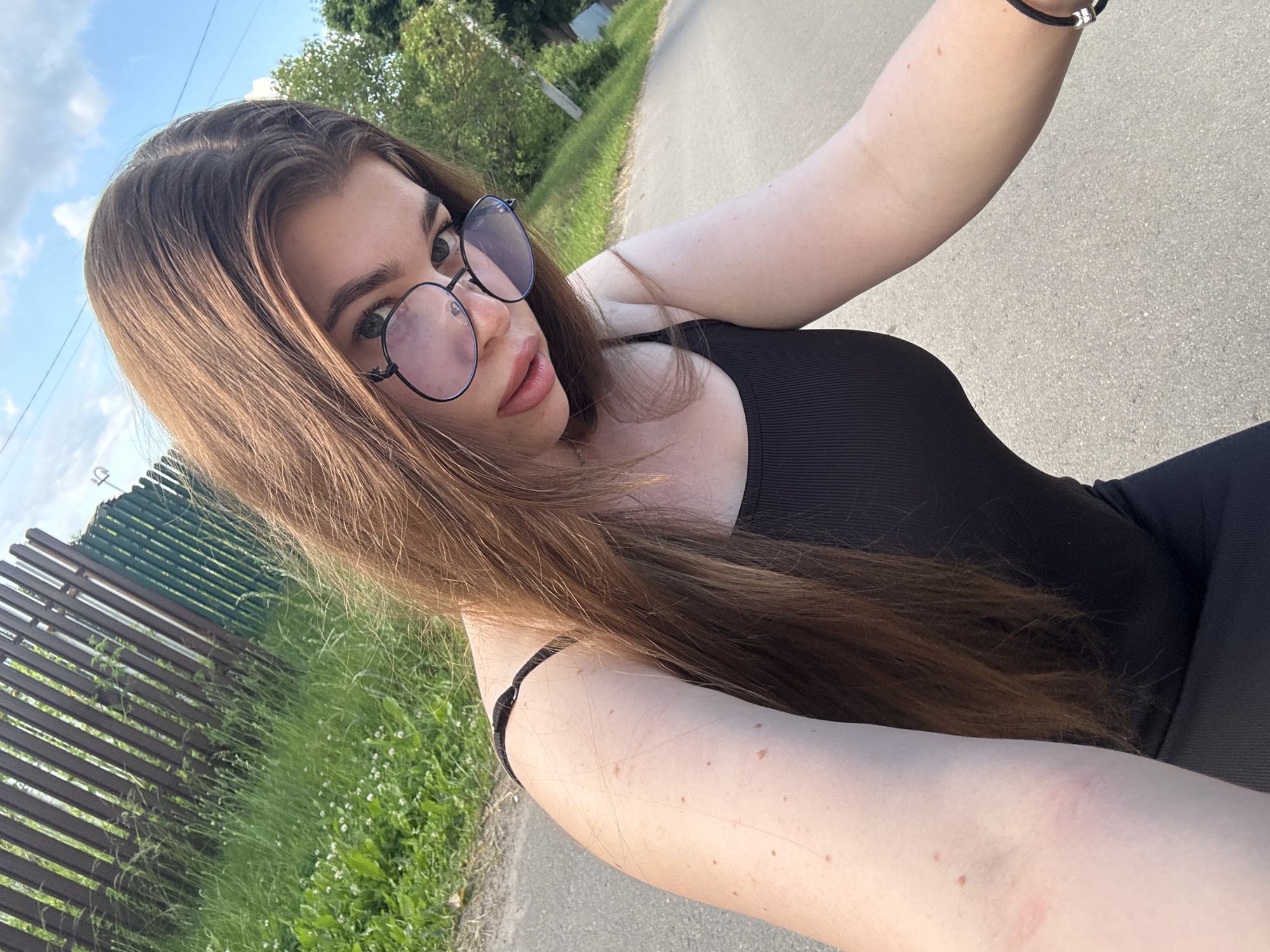 A young woman with long, brown hair and glasses takes a selfie outdoors. She is wearing a black tank top. The background shows a wooden fence, green grass, and a paved road. The sky is visible with scattered clouds.