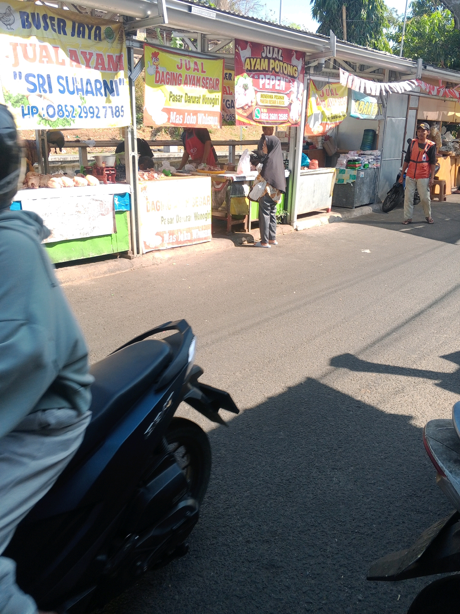 The image captures a bustling street scene in a market area of Wonogiri, Indonesia, likely during daylight hours given the bright sunlight and strong shadows. In the foreground, a dark blue motorcycle is partially visible from the perspective of someone riding it, with the rider's light gray jacket and the motorcycle's seat and rear fender in view. The road surface is asphalt, and a large shadow from the motorcycle and rider stretches across it.
The background features several market stalls with colorful banners advertising the sale of chickens. One prominent banner, yellow with blue and red text, reads "BUSTER JAYA JUAL AYAM 'SRI SUHARNI' HP: 0852-2992 7785". Adjacent to it, another banner in yellow and red proclaims "JUAL DAGING AYAM SEGAR Pasar Darurat Wonogiri Mas Joko Whispu". Further down, more stalls are visible with various goods, including what appear to be packaged items and possibly a refrigerator unit.
Several people are present. A vendor, wearing a red vest over a light-colored shirt and pants, stands near a stall, looking towards the right. A woman wearing a black hijab and a patterned shawl is interacting with someone at a stall, holding a plastic bag. Another person, partially obscured, is also seen behind a stall. The architectural style suggests a typical open-air market with simple structures and awnings. The overall impression is one of a lively commercial environment.