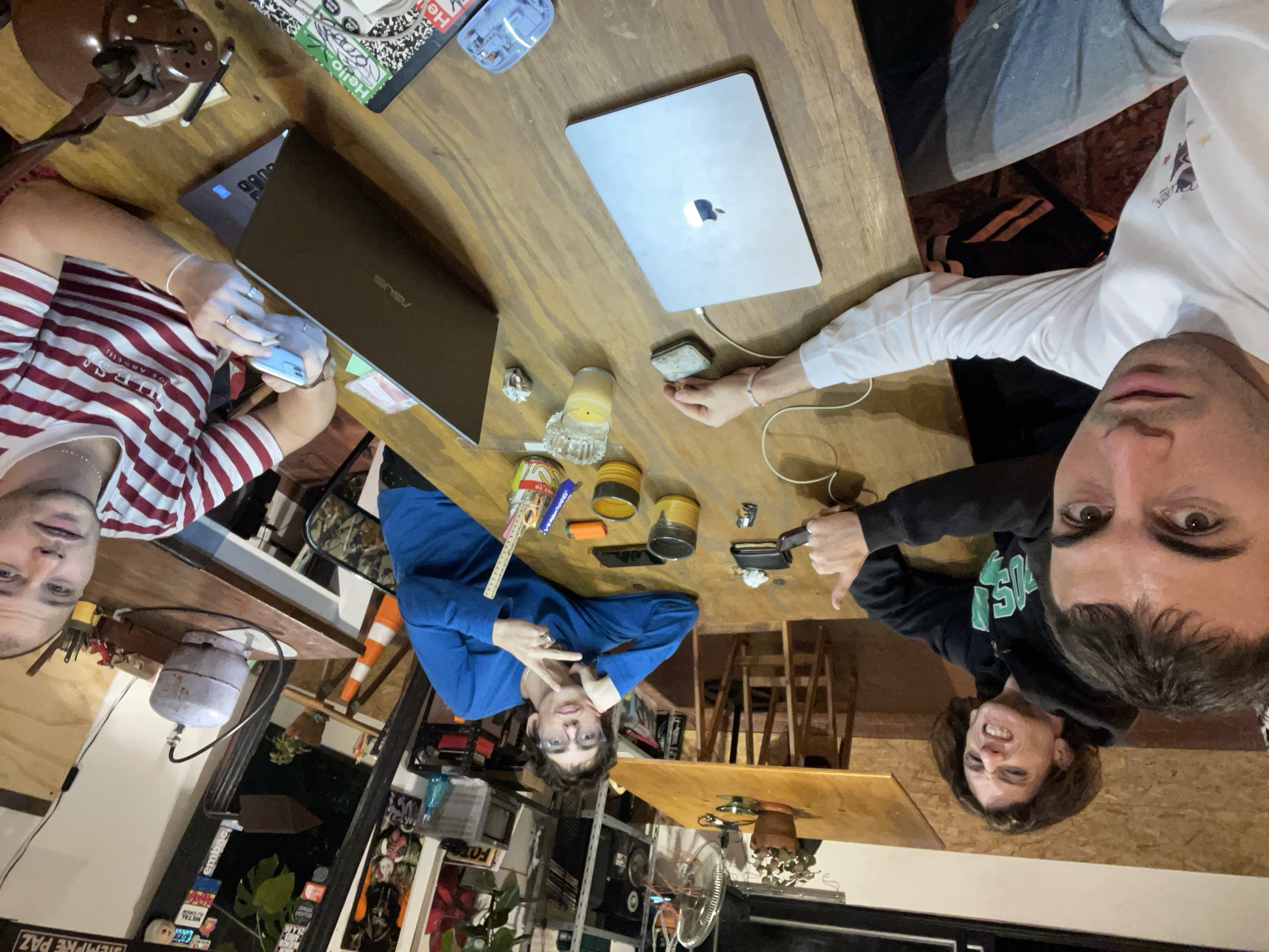 A wide-angle, casual group photo, rotated 90 degrees clockwise, captures four young men gathered around a large wooden table in an indoor setting, likely in Buenos Aires, Argentina. The scene appears to be taking place in the evening or night, illuminated by artificial light and a burning candle.
On the left, a man wearing a red and white striped "GUESS" t-shirt holds a light-blue phone or rolled cigarette, looking directly at the camera. An open ASUS laptop and a brown table lamp are next to him. Opposite, a man in a blue long-sleeved top, wearing glasses, makes a peace sign with both hands, also engaging with the camera. Nearby items include a ruler, a lighter, and small metal cans.
On the right, a person's arm is visible next to a charging MacBook with a glowing Apple logo. Further right, a man in a black hoodie with partially visible green text, possibly "WISCONSIN," gives a playful thumbs-down gesture while looking at the camera. The face of a fourth man, with dark hair, is very close to the right edge of the frame, suggesting he might be taking the photo.
The cluttered wooden table holds various items: two laptops, a lit candle in an ashtray, a blue water bottle, and other small objects. The background reveals a busy environment with shelves filled with plants, books, and miscellaneous items, alongside a fan, a small white gas cylinder, and an orange cone. Visible text includes "GUESS" on the striped shirt, "ASUS" on a laptop, "Hello" on a sticker, and "SIEMPRE PAZ" on a black and white sticker in the bottom left. The overall atmosphere is relaxed and social.