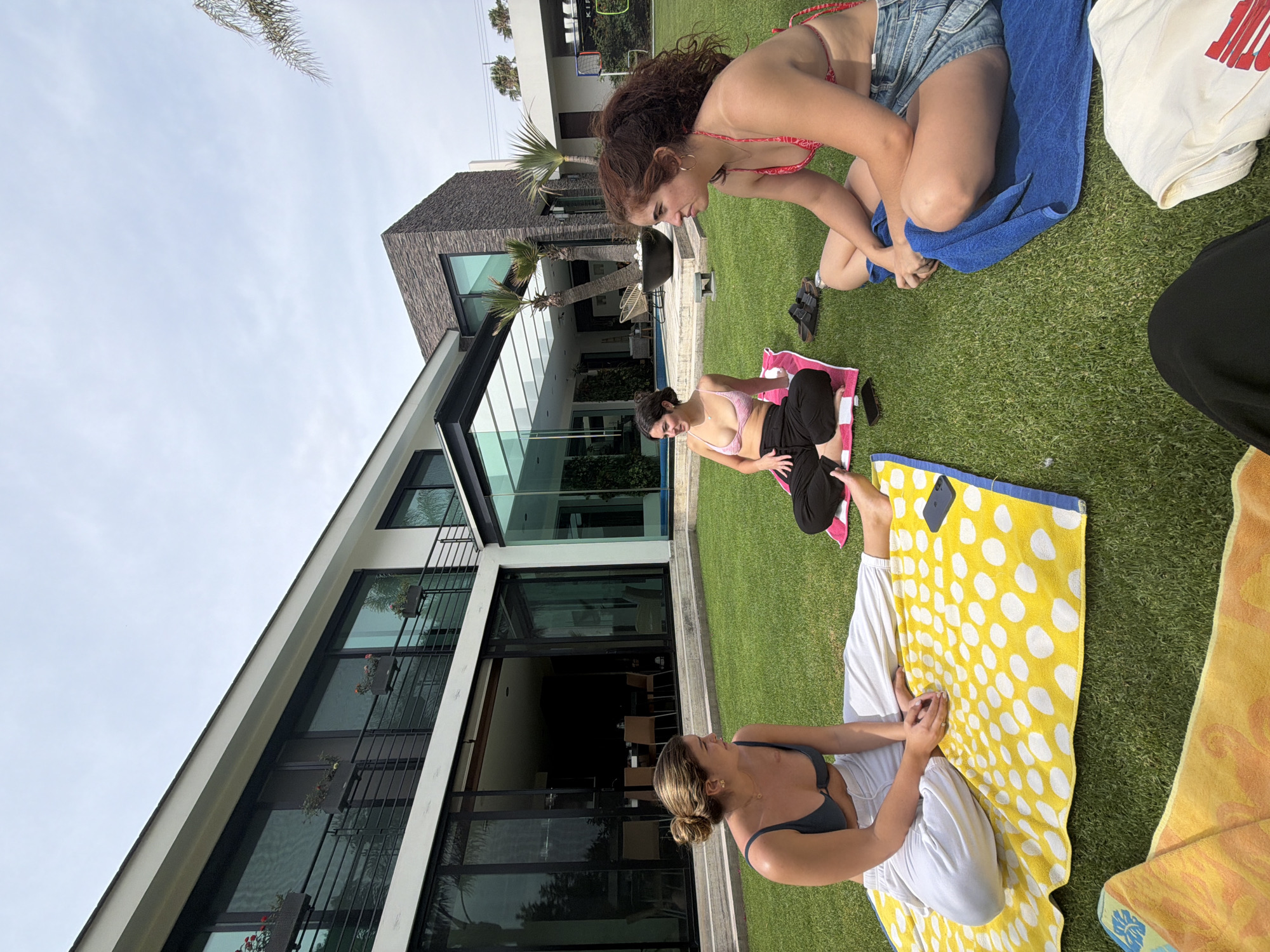 The image captures an outdoor scene on a grassy lawn. Three women are present: one kneeling on a blue towel, wearing a red and white bikini top and denim shorts; another seated on a pink towel, wearing a pink and white striped bikini top and dark pants; and the third also seated, on a yellow and white polka-dot towel, wearing a dark top and white pants. Various items such as towels of different colors and patterns, clothing, and a mobile phone are scattered around the lawn. A modern house with large glass windows and a gray stone structure is visible in the background. The location appears to be a sunny outdoor area, possibly a backyard or garden of the house.