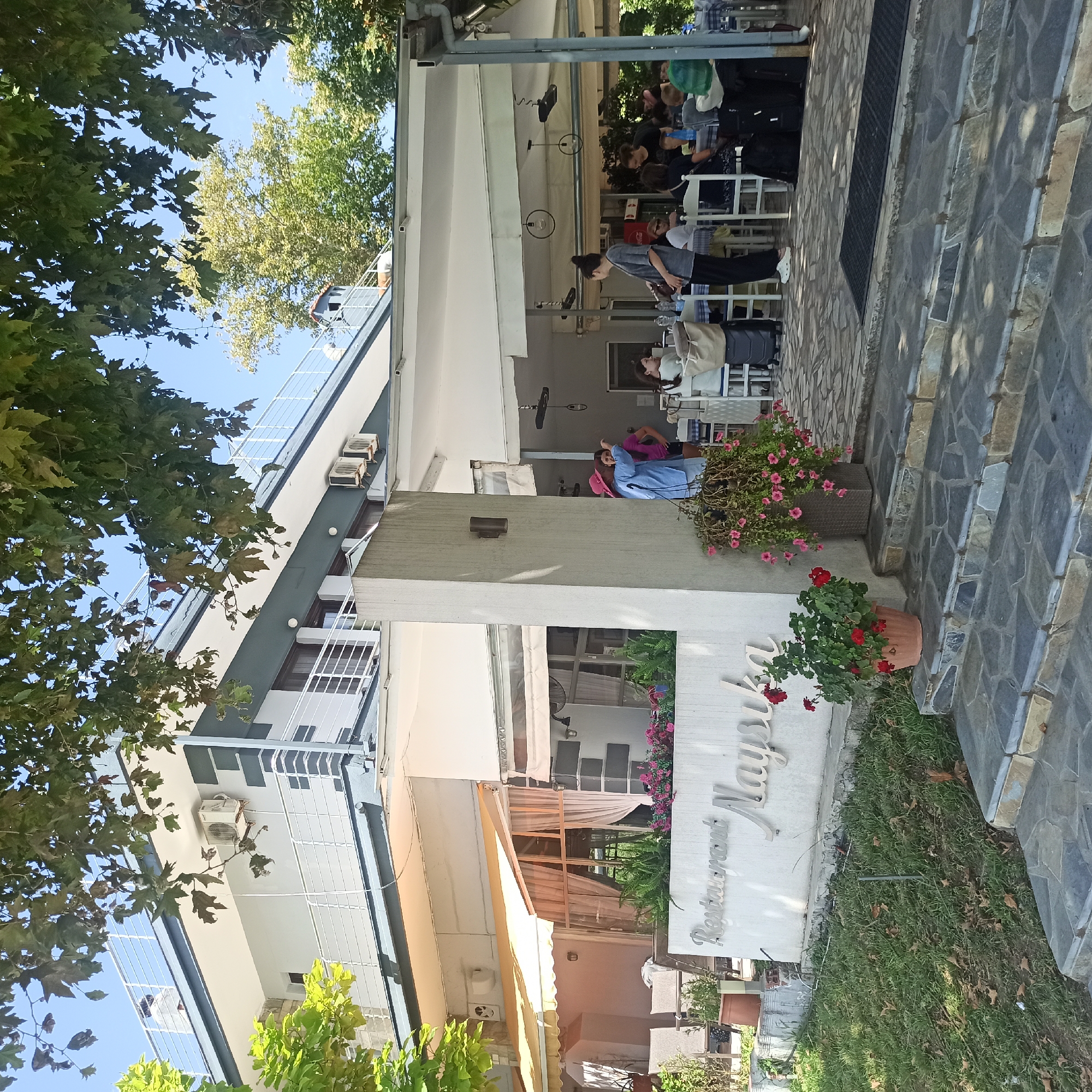 The image captures the exterior of a two-story building with a sign that reads "Restaurant Naysika" in Limani Litochorou, Greece. The building has white walls, dark gray accents, and balconies with white railings. An outdoor seating area for the restaurant is visible, covered by awnings, with tables and chairs set up. Several people are seated at the tables, engaged in dining or conversation. A woman in a pink hat and blue shirt is standing near a concrete pillar, possibly interacting with someone. The area is surrounded by greenery, including trees and potted plants with pink and red flowers, and the ground is paved with stone. Steps lead up to the restaurant entrance. The sky is clear and blue, indicating a sunny day. Luggage, including suitcases, is visible near some of the tables, suggesting that the establishment may also cater to travelers.