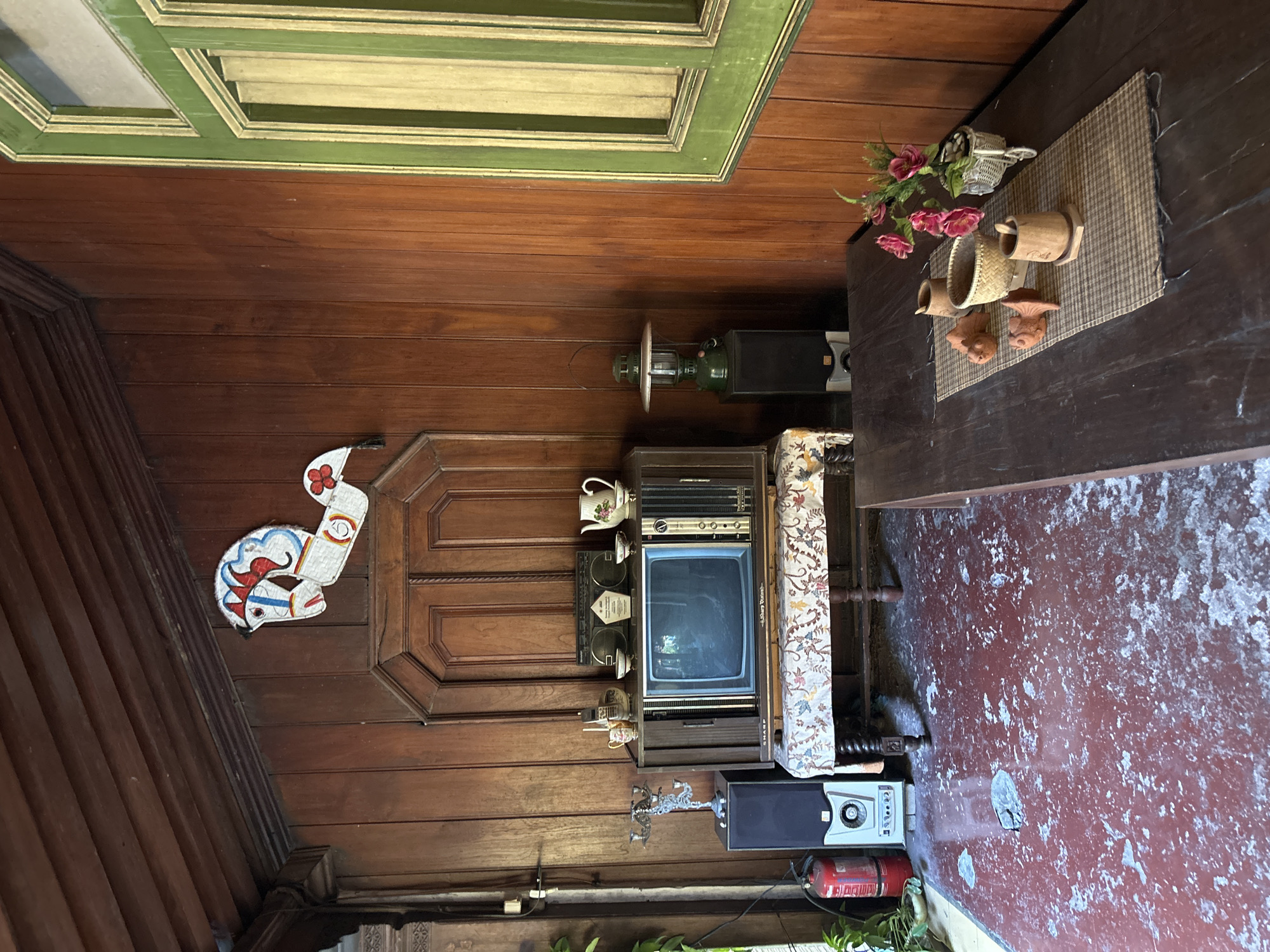 The image depicts an interior scene, possibly a veranda or a living area, with predominantly wooden elements and a rustic aesthetic. The walls and ceiling are paneled with dark wood, creating a warm and enclosed atmosphere. In the center of the image, a vintage television set sits on a patterned cloth-covered table. Above the television, a decorative wooden cutout of a horse with red and blue accents is affixed to the wall.
To the left of the television, a black speaker is placed next to a red fire extinguisher. To the right of the television, another black speaker supports a green kerosene lantern. A vintage radio-cassette player is perched on top of the television cabinet, along with a white teapot and several small bowls.
The floor is a distinctive red with patches of what appears to be peeling paint or concrete, giving it a distressed look. In the foreground, a dark wooden table is set with a woven placemat. On the placemat are decorative wooden fish figures, a small basket, and small wooden cups. A small bouquet of pink flowers in a miniature bicycle basket adds a touch of color.
A green-framed window is visible on the right side of the image. The overall impression is one of a nostalgic and somewhat cluttered space, possibly a family heirloom or a space designed to evoke a sense of the past. The mention of Banguntapan, Indonesia, suggests a specific geographical context, hinting that this scene might be representative of traditional Indonesian home decor or a specific establishment within that locale.