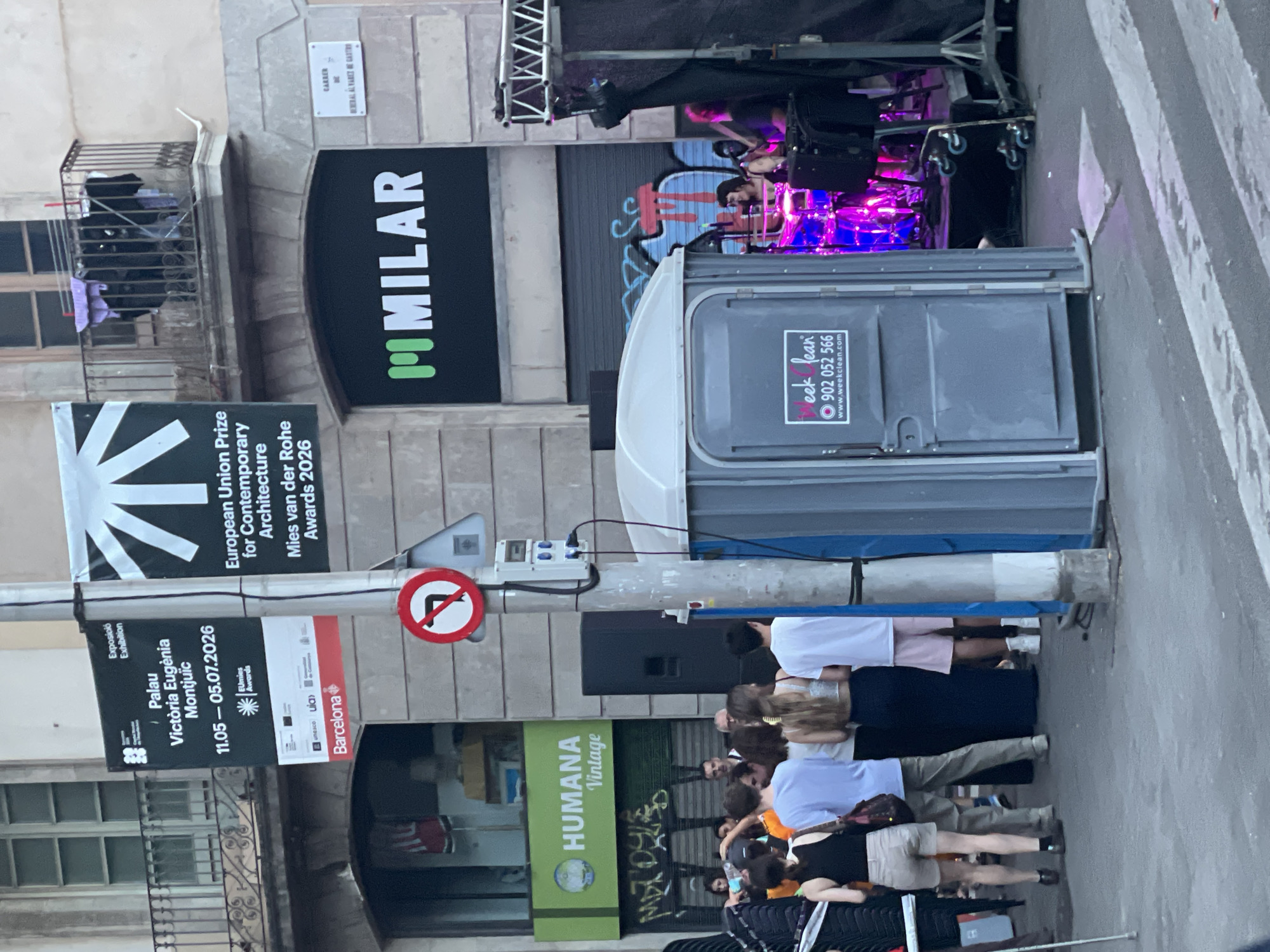 The image shows a street scene in Barcelona, Spain, with a band performing on a stage on the right. In the foreground, there is a portable toilet with a sticker for "Week Clean". A group of people are standing and walking on the street. Above the street, there are buildings with balconies and posters advertising exhibitions. One poster is for "Palau Victòria Eugènia Montjuïc" and another is for "European Union Prize for Contemporary Architecture". A street sign is visible on the left with "HUMANA Vintage" written on it. The overall scene suggests an outdoor event or festival taking place in the city.