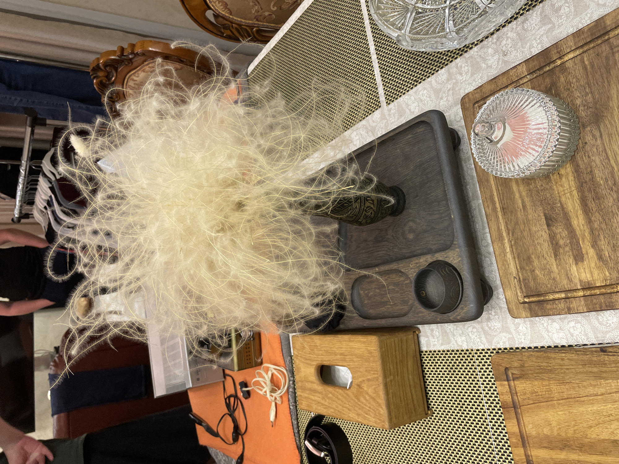 A slightly high-angle shot shows a collection of objects arranged on a surface with a patterned placemat, suggesting an interior setting. The most prominent feature is a large arrangement of feathery plants, likely decorative, emanating from a vase on a dark, possibly wooden, tray with two small circular compartments. To the right of the tray, there are two wooden cutting boards and a clear glass container with a ribbed texture and a lid, revealing pink contents inside. To the lower left, an orange surface is visible with audio cables and connectors, along with a wooden tissue box. The lower part of the frame reveals what looks like a part of a black belt with a silver buckle. The surface appears to be a table, covered with the patterned placemat. Beyond the objects, there are indistinct figures, clothing racks, and what appears to be the back of a chair with ornamental wood carvings, indicating a domestic or commercial space, not necessarily a landscape consistent with Лесной Пейзаж Кп (шарапово), Russia. The scene is static and does not indicate any specific action.