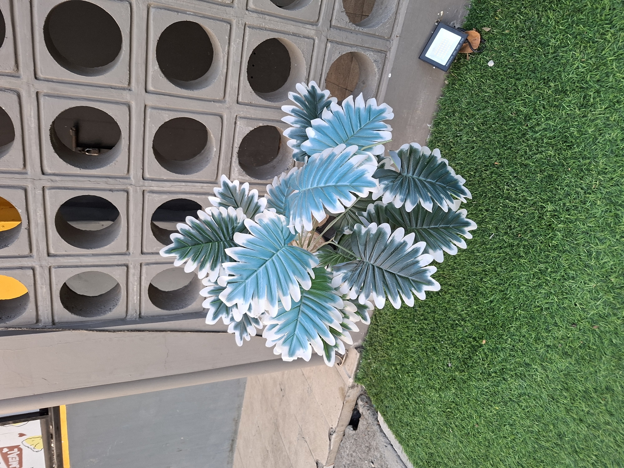 A close-up, high-angle shot captures a cluster of artificial houseplants with vibrant teal and white leaves, placed on a patch of artificial turf. The plants are positioned in front of a grey, perforated concrete wall with square openings. To the left of the plants, a grey concrete sidewalk is visible, cracked and worn. A small black floodlight with a white panel sits on the artificial turf near the bottom right corner of the frame. The scene appears to be an outdoor area, possibly a public space or a business facade in Surawangi, Indonesia, with the style of the wall and the presence of artificial plants suggesting an urban setting. No people are present in the image.
