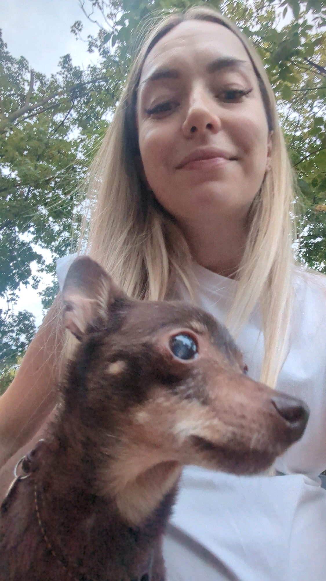 A low-angle, selfie-style photograph captures a woman and a small dog outdoors. The woman, with blonde hair and visible makeup, is looking down and smiling slightly. She is wearing a white t-shirt. The dog, a small brown breed, is in the foreground, with its head turned to the side and its eye visible. The background consists of leafy green trees and a cloudy sky, suggesting an outdoor setting. The image does not contain any discernible information that would allow for identification of a specific location in Chișinău, Moldova.