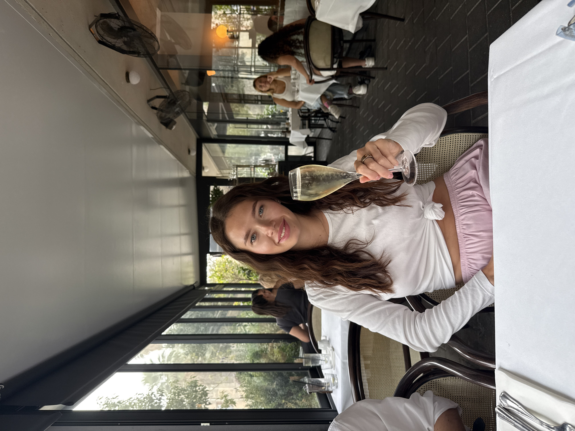 This medium-shot photograph, taken in a bright restaurant setting in Tel-Aviv, Israel, features a young woman smiling directly at the viewer. She is positioned slightly off-center, holding a champagne flute containing a pale, effervescent liquid. Her long, dark brown hair flows over her shoulders, framing her face and blue eyes. She is wearing a white, long-sleeved crop top, knotted at the midriff, paired with light pink, satin-like elasticated trousers. A dark ring adorns her left ring finger.
She is seated at a table covered with a white tablecloth, with a section of the table and wrapped silverware visible in the bottom right corner. The restaurant's interior design includes dark, patterned floor tiles and rattan-backed chairs with dark wooden frames. Overhead, ceiling fans and light fixtures are mounted on a light-colored ceiling.
Large, expansive glass windows, framed in black, form the left side of the restaurant, offering a view of outdoor greenery and allowing ample natural light to illuminate the space, indicating daytime. Reflections in these windows show other patrons, including another young woman seated at a table in the background, also appearing to smile. The overall impression is one of a relaxed, social gathering or meal in a stylish, well-lit establishment, with the woman's joyful expression conveying a pleasant and enjoyable experience.