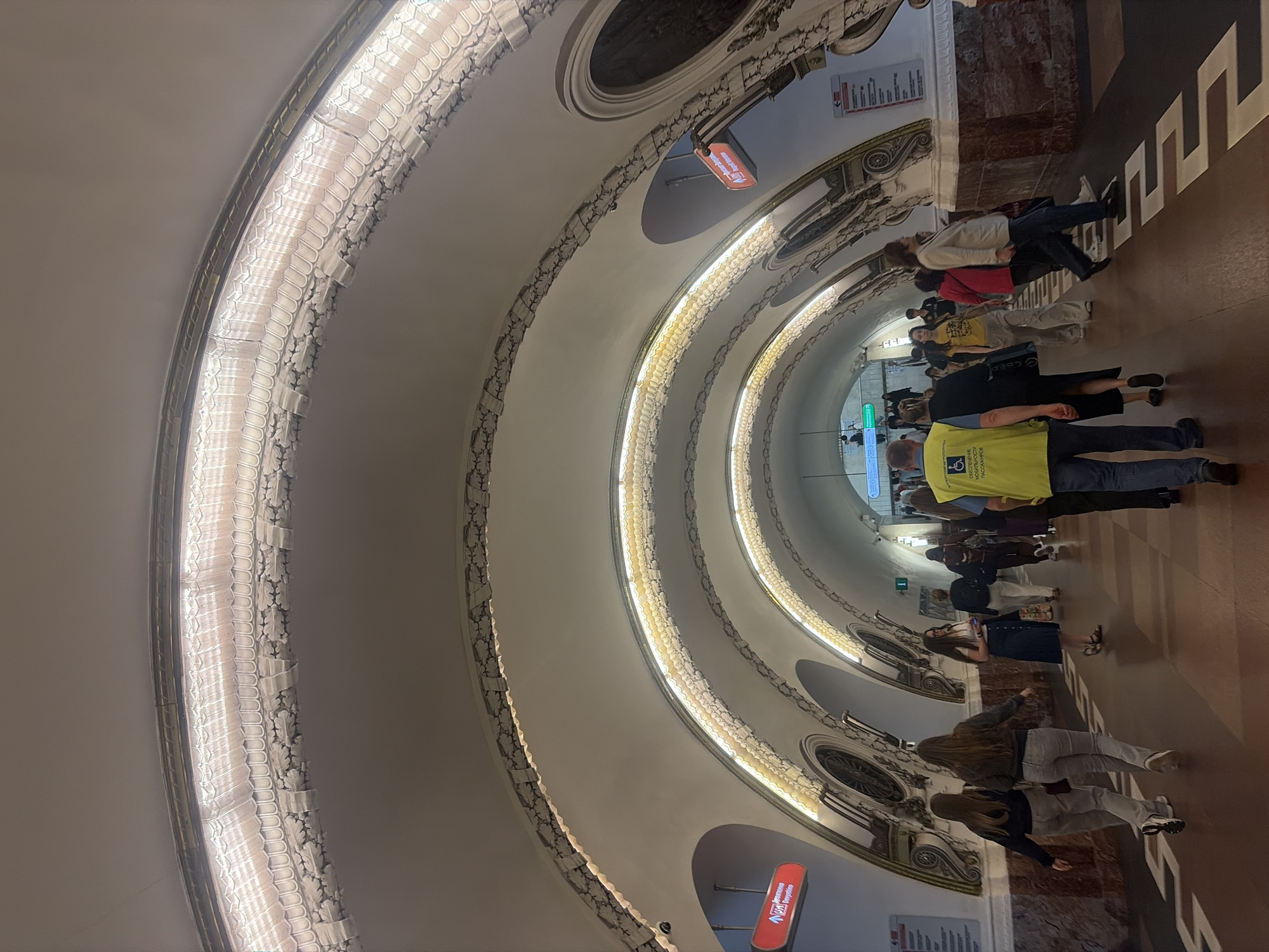 The image shows a curved interior with ornate architecture, likely a metro station in Saint Petersburg, Russia, given the context. A group of people are walking down a corridor, suggesting they are commuters or tourists. The lighting is dim, with bright, decorative lights lining the curved walls, giving the scene an illuminated yet slightly dramatic feel. The architecture features a repeating pattern of arches and decorative moldings, with some areas of marbled or dark stone. The floor is a polished dark wood with a geometric pattern in a lighter color.
Several people are visible, most are walking away from the camera, appearing to be in motion. One person in the foreground wears a bright yellow vest with text on it. Another person further down the corridor wears a black t-shirt with yellow text. There are also signs visible on the walls, one red illuminated sign has Cyrillic text. The overall impression is one of a busy, yet grand, public space. The orientation of the image is rotated, with people appearing to walk up the image.