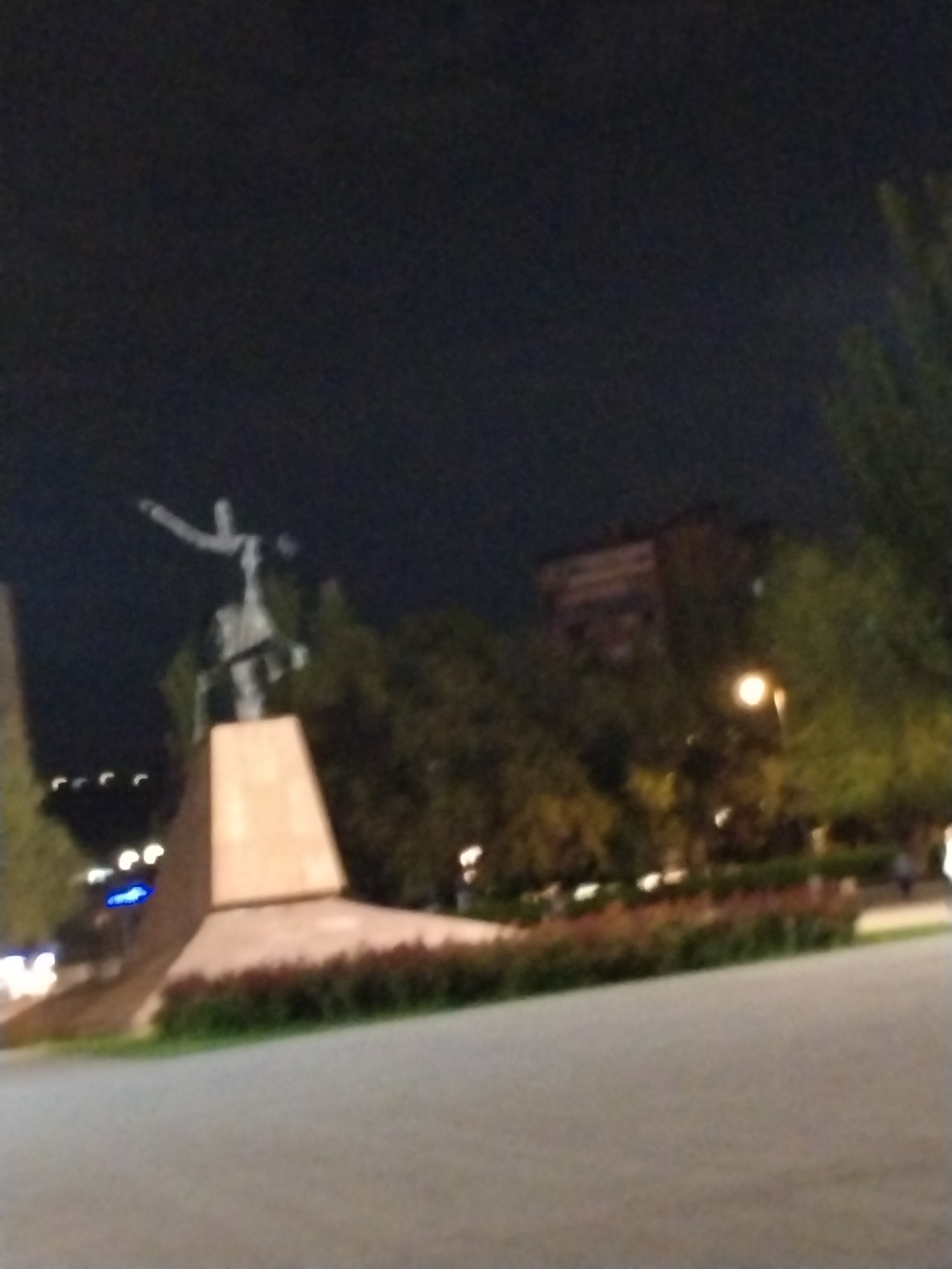 This nighttime photograph captures a prominent statue, likely the Mother Armenia monument, situated in Yerevan, Armenia. The central subject is a large, dark-colored statue of a figure, appearing to be female, with one arm outstretched or holding an object, standing on a massive, angular, light-colored stone pedestal. The statue is partially illuminated, standing starkly against a very dark, moonless sky.
The scene is an outdoor public space, possibly a park or square. Surrounding the statue and extending into the mid-ground are numerous trees with dark foliage, which appear green under artificial lights. A large building with visible windows can be vaguely discerned through the trees in the right background. Blurry points of light, consistent with distant city lights or streetlights, dot the background and foreground, creating a soft glow.
In the foreground, a wide, paved area, likely a walkway or plaza, occupies the bottom of the frame. This paved surface is bordered by a low, well-maintained strip of reddish-green shrubbery. The overall ambiance is tranquil and monumental, with no specific activity or people clearly visible. The time of day is undeniably night, illuminated solely by artificial light sources. No visible text can be identified in the image. The photograph has a slight blur throughout, suggesting movement or a lower light setting during capture.