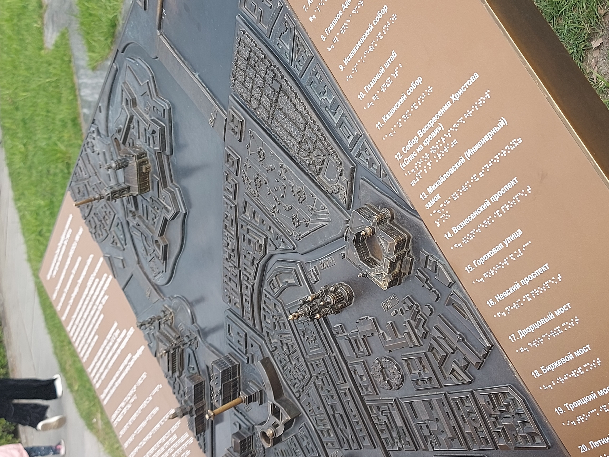This image shows a tactile city map of Saint Petersburg, Russia. The map is made of a dark gray metal and features raised representations of buildings and streets, allowing visually impaired individuals to understand the city's layout. Braille and printed text labels key landmarks, including Nevsky Prospekt, Kazan Cathedral, The Church of the Savior on Spilled Blood, and several bridges. The map is outdoors and partially surrounded by grass. People's legs and feet are visible to the left of the map, indicating its accessibility.