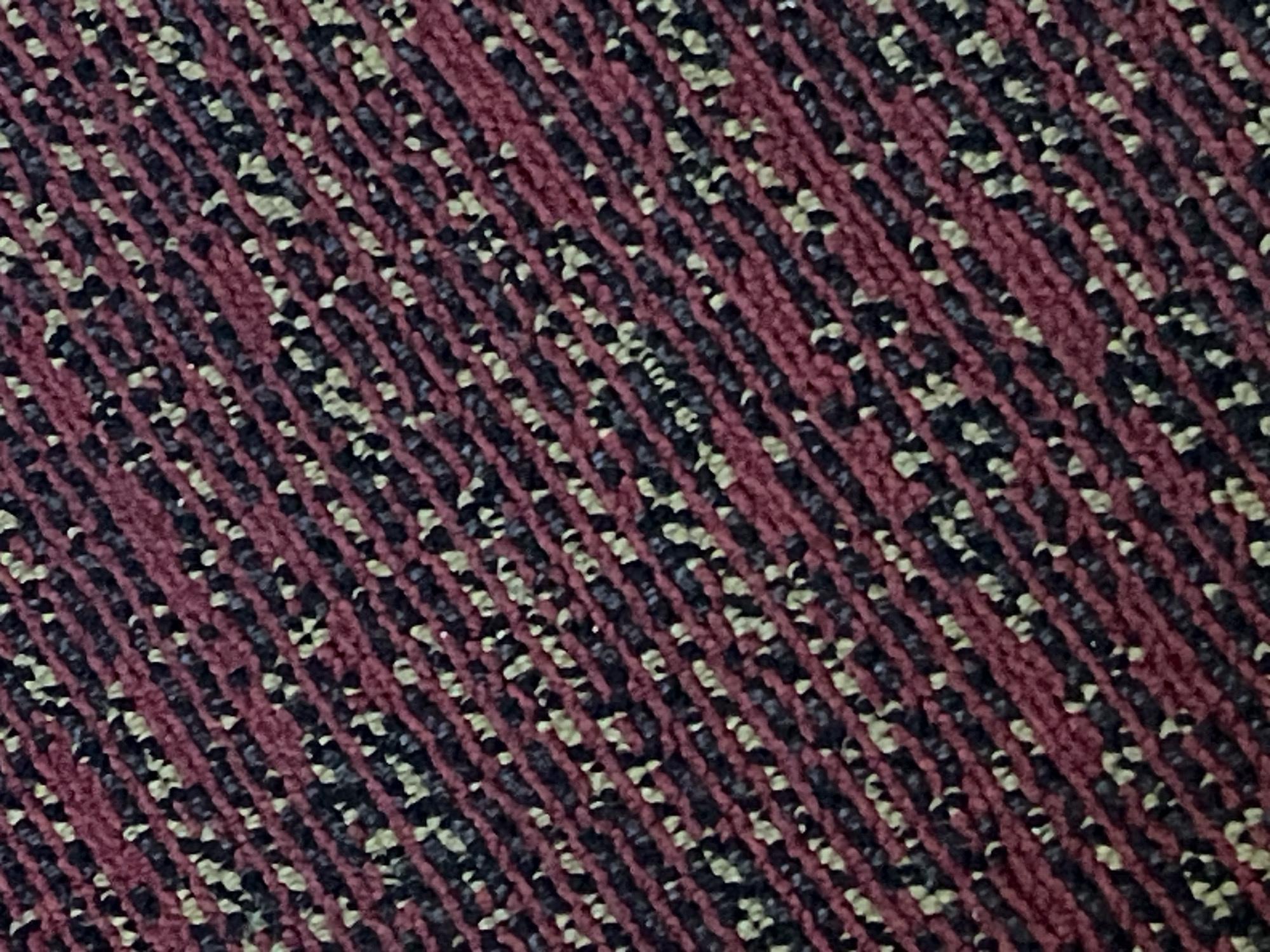 The image displays a close-up, angled view of a carpet with a diagonal striped pattern. The stripes are composed of dark red, black, and small specks of light yellow or off-white. The texture appears to be looped or cut pile, typical of commercial carpeting. There are no people, objects, or discernible actions visible in the image. The scene is a flat, textural surface. The image does not provide any information about its location in Bac Cam Ranh Ward, Vietnam, or any other geographical context.