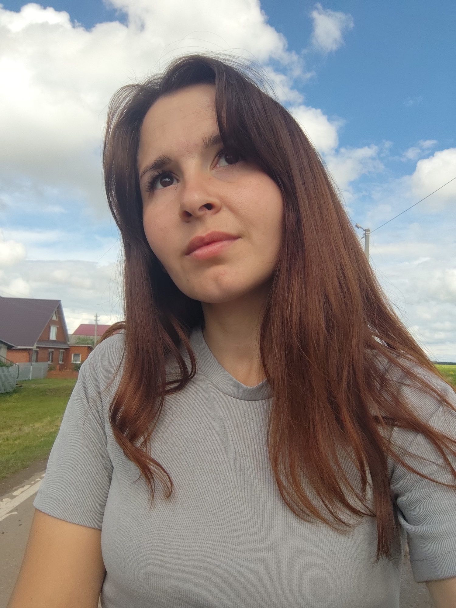 A medium close-up outdoor portrait captures a young woman in a rural or village setting, identified as Фоки, Russia. She has long, wavy brown hair and is wearing a light grey, ribbed short-sleeved t-shirt. Her head is tilted slightly, and she looks upwards and to her left with a neutral, thoughtful expression. Her eyes are dark brown, and her lips appear lightly colored.
The background features a bright, partly cloudy sky with patches of vivid blue. On the left side, two houses with dark brown roofs and brick walls are visible, surrounded by green grass and a fence. A paved or dirt road runs along the lower left of the frame. To the right, a utility pole with overhead wires is present, and a distant green field stretches under the horizon. The scene takes place during daylight hours, indicating bright weather conditions with scattered clouds. There is no visible text in the image.