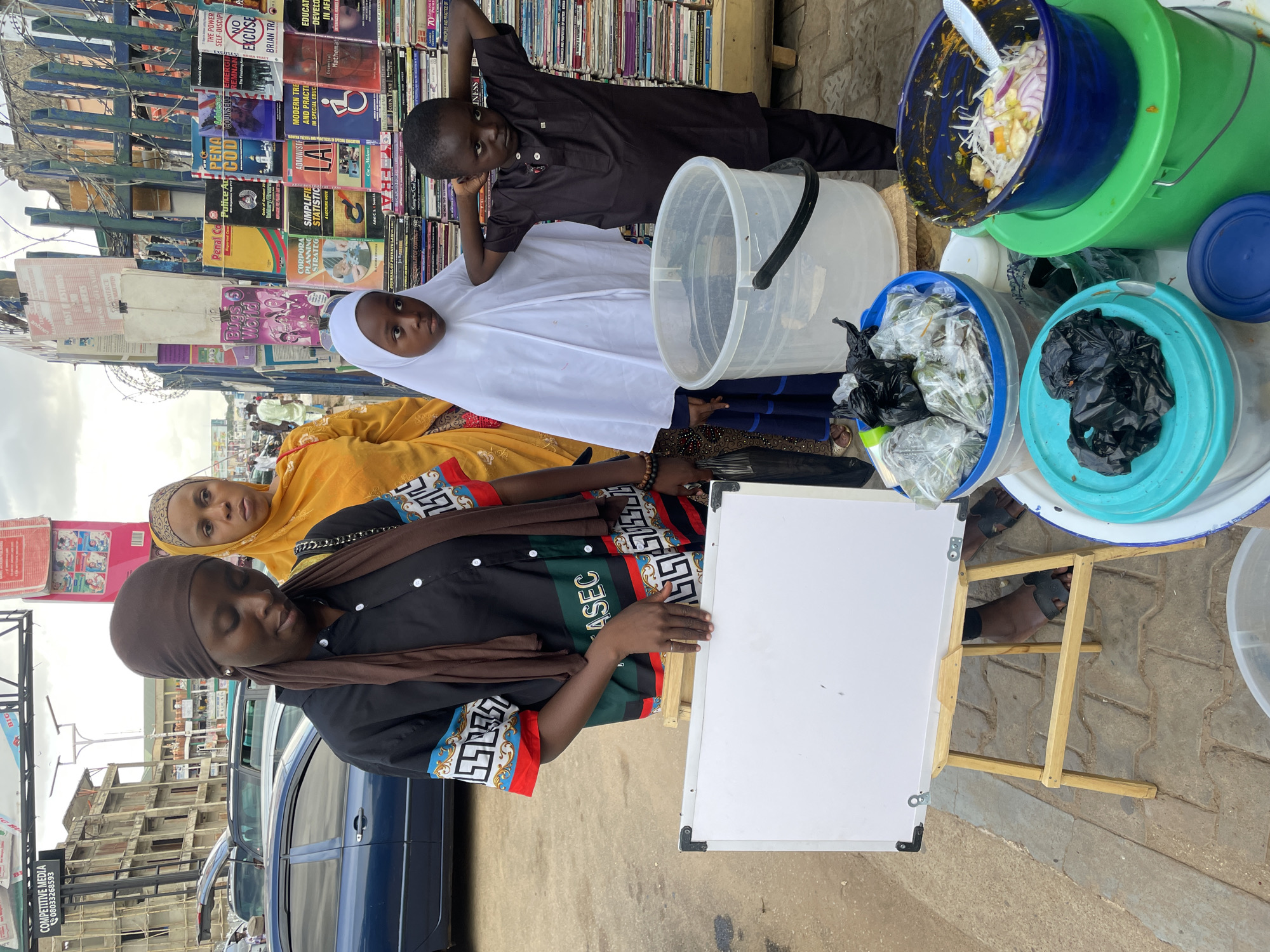 Here is a detailed description of the image:
**Scene:**
The image captures a street scene in what appears to be Jos, Nigeria. It shows a roadside vending setup. A table, made with a white board and wooden legs, is the central focus.
**People:**
* Three females are present, they all appear to be of Nigerian descent. The main focus is the woman standing closest to the table, wearing a black shirt with a brown scarf and colorful printed edges.
* Behind her, another woman wears a yellow hijab and looks at the camera.
* Two children are present. One is standing behind the table in a white hijab. The other one is resting his head on a book.
* Additional pedestrians are visible in the background.
**Objects:**
* A wide assortment of used books is displayed on a metal shelf behind the vendor. The titles are in English and appear to cover a range of topics, including law, business, education, and self-help.
* Several buckets of food items are placed on the table. They contain salad and other ingredients.
* A white board propped up by a wooden table.
* A blue car is parked on the road.
**Actions and Interactions:**
* The main vendor is standing at the table.
* The scene suggests the preparation and sale of food to passersby.
**Environment:**
* The setting is an urban street.
* A building is partially visible in the background.
* The sky is cloudy.
* The overall environment suggests a bustling street market in an urban environment.