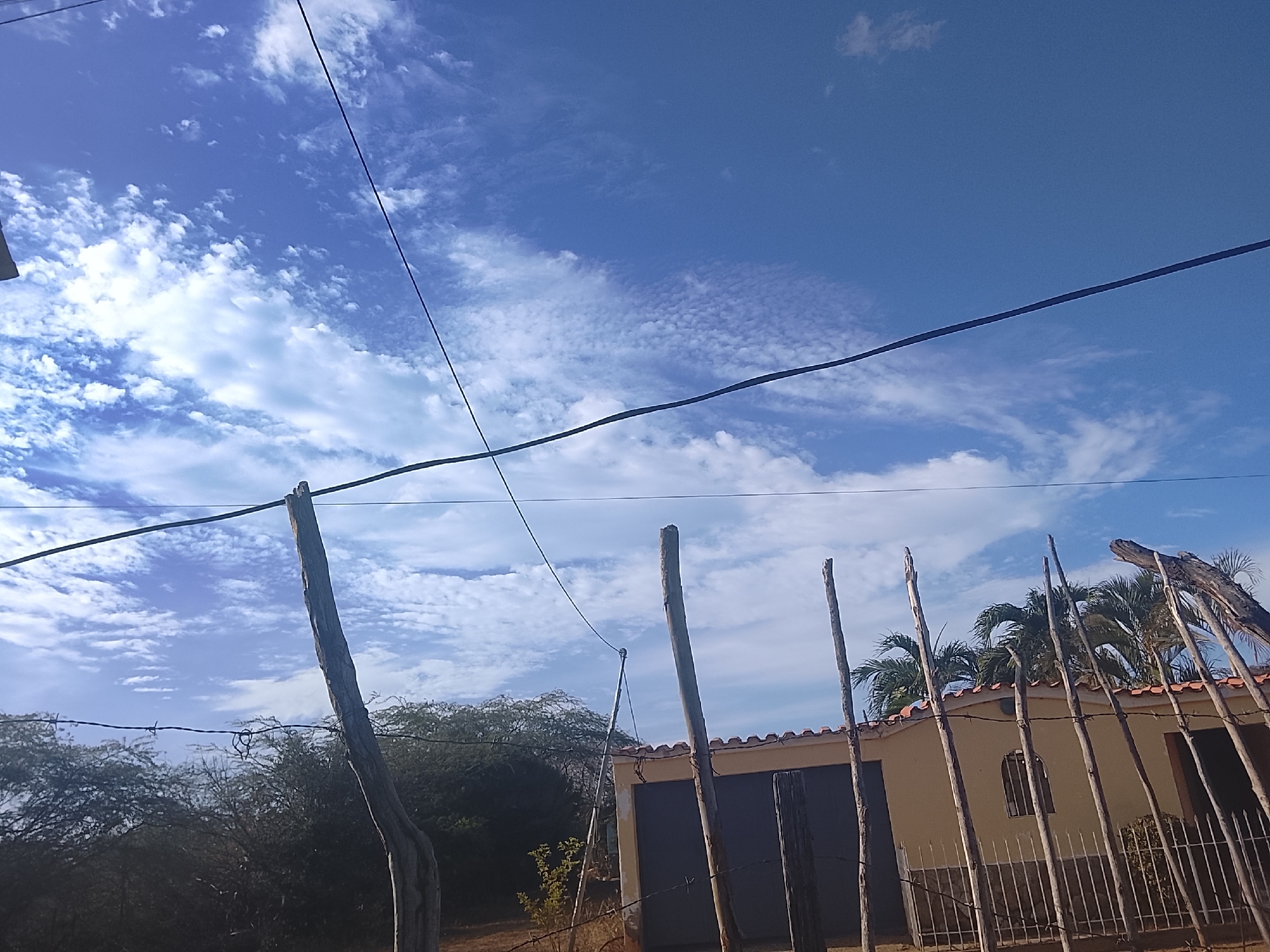 The image depicts a daytime scene in what appears to be a rural area. The sky is blue with scattered clouds. In the foreground, there are wooden fence posts with barbed wire running along them. Behind the fence, there is a small, beige-colored building with a red tiled roof. Vegetation, including trees and palms, is visible in the background. Overhead, power lines cross the sky. There is nothing to indicate this location being in Altagracia, Venezuela. No people are visible in the image.