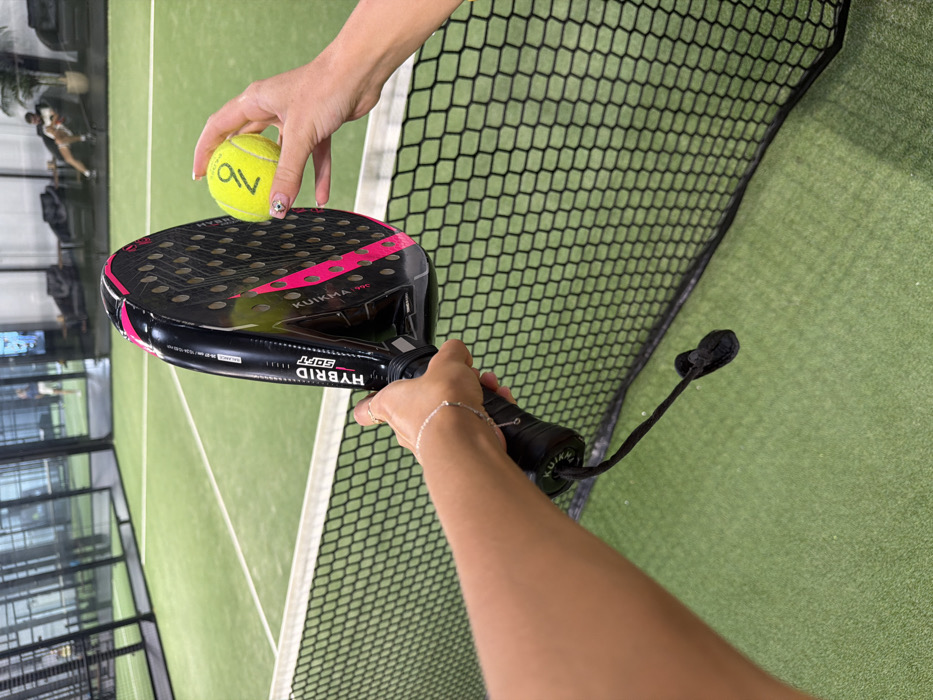 The scene is observed from a first-person perspective within an indoor padel court facility in Choeng Thale, Thailand. In the immediate foreground, a person's left hand grips the black handle of a black and pink Kuikma Pro Hybrid Soft padel racket; a black wrist strap hangs from the handle, and a silver bracelet is visible on the wrist. Concurrently, a person's right hand holds a fluorescent yellow padel ball, featuring a black "Z" logo; this hand displays manicured nails with a discernible design. Both hands are positioned just above a black, hexagonal-mesh padel net that stretches across the mid-ground. The background reveals a green artificial turf padel court with white demarcation lines, enclosed by dark mesh fencing. Beyond the court, another individual is seen standing in a light-colored shirt on an adjacent court area to the left. To the right, two additional individuals are seated on dark lounge furniture in a designated spectator or rest area. The facility appears modern, characterized by dark interior elements and large glass panels in the distant background.