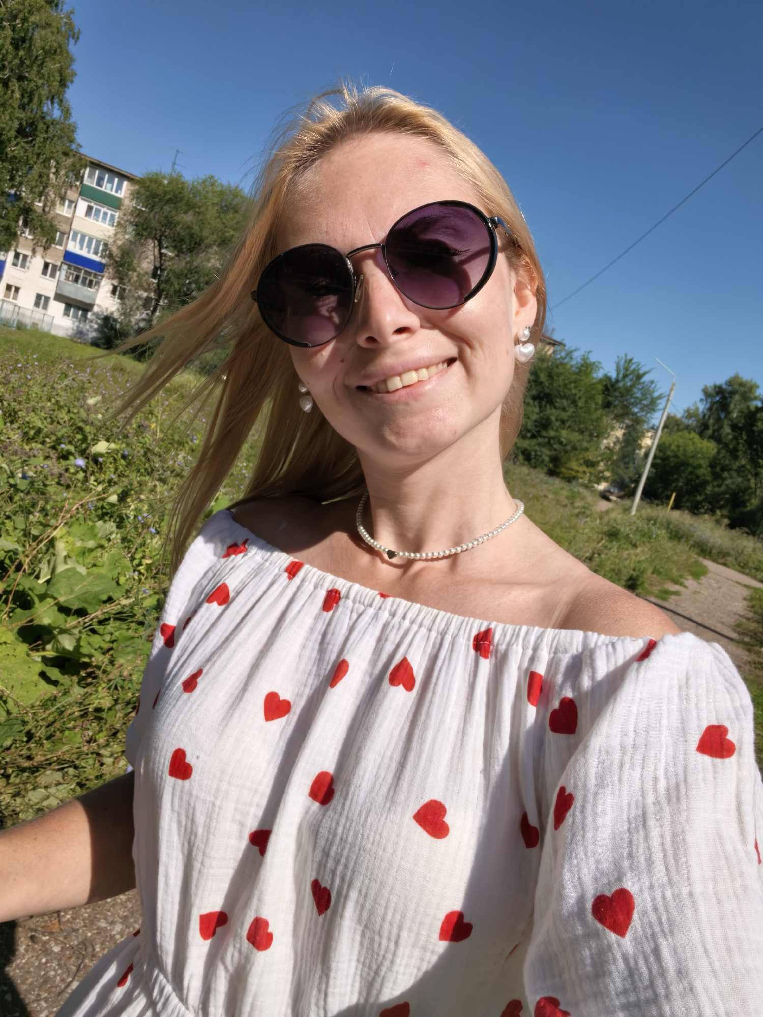 A woman is taking a selfie outdoors in Sterlitamak, Russia. She has blonde hair and is wearing large, round sunglasses, pearl earrings shaped like hearts, and a white off-the-shoulder top with red heart patterns. She is also wearing a pearl necklace with a small heart pendant. The background shows a blue sky, trees, a building, and vegetation. The lighting suggests it is daytime, likely sunny.