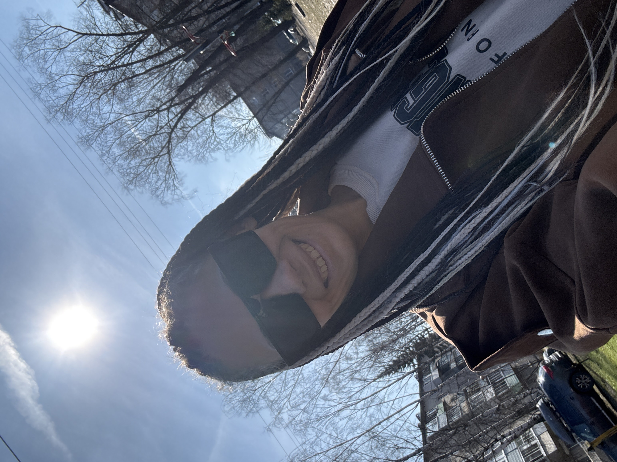 A woman with dark braids and sunglasses is seen from a low angle, smiling. She is wearing a brown jacket over a white shirt with black lettering. In the background, a blue sky with a bright sun is visible, as well as bare tree branches. There is a multi-story building in the distance, along with a parked blue car and a patch of green grass. The location is a city street of Unknown, Unknown.