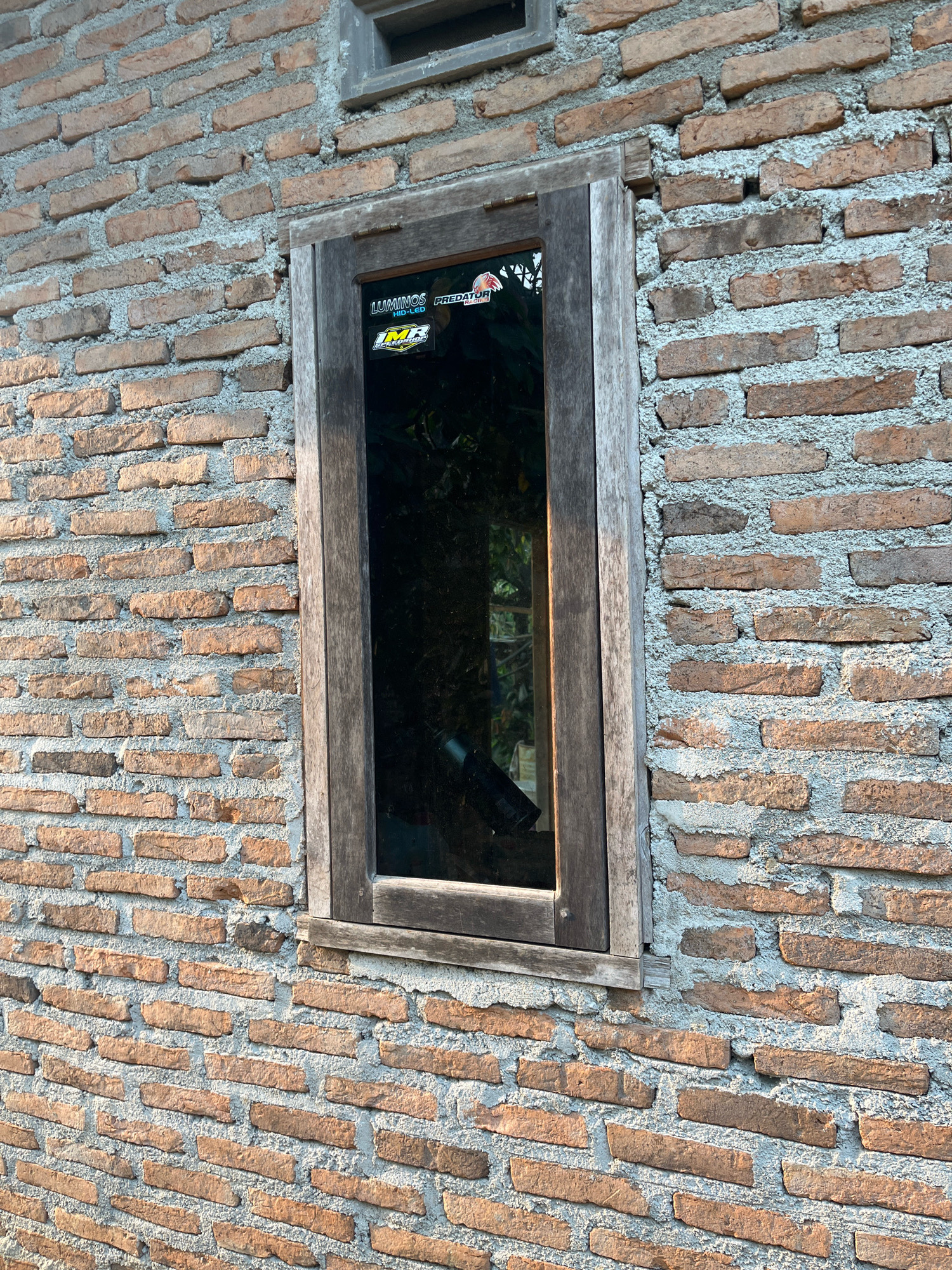 This is an exterior, eye-level shot of a window set into a brick wall in Kebumen, Indonesia. The wall is constructed of roughly uniform, reddish-brown bricks laid in a horizontal pattern with gray mortar. The window frame is made of weathered, light-brown wood. The glass in the window is dark and reflective, obscuring the interior. Two stickers are visible on the upper right portion of the windowpane. The upper sticker has the word "LUMINOS" in large white letters above "HID-LED" in smaller white letters, with a black graphic of a stylized eye. Below that, a second sticker is mostly yellow and black, featuring an emblem with the letters "IMR" in large black font. The lighting suggests late afternoon or early morning, with warm light casting shadows across the brickwork. The overall scene depicts a humble, possibly rural or older, structure.