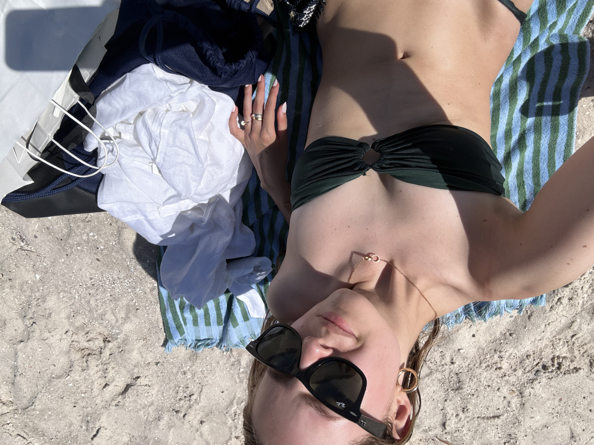 A woman is lying on a beach towel on the sand, taking a selfie. She is wearing a dark green bikini top and sunglasses. She has on hoop earrings and a necklace. To her right are some clothes and a white paper bag. The sand appears to be light in color, and there is some debris on it. The sun is shining brightly, casting shadows on the scene. The location is the city of Miami, United States of America.