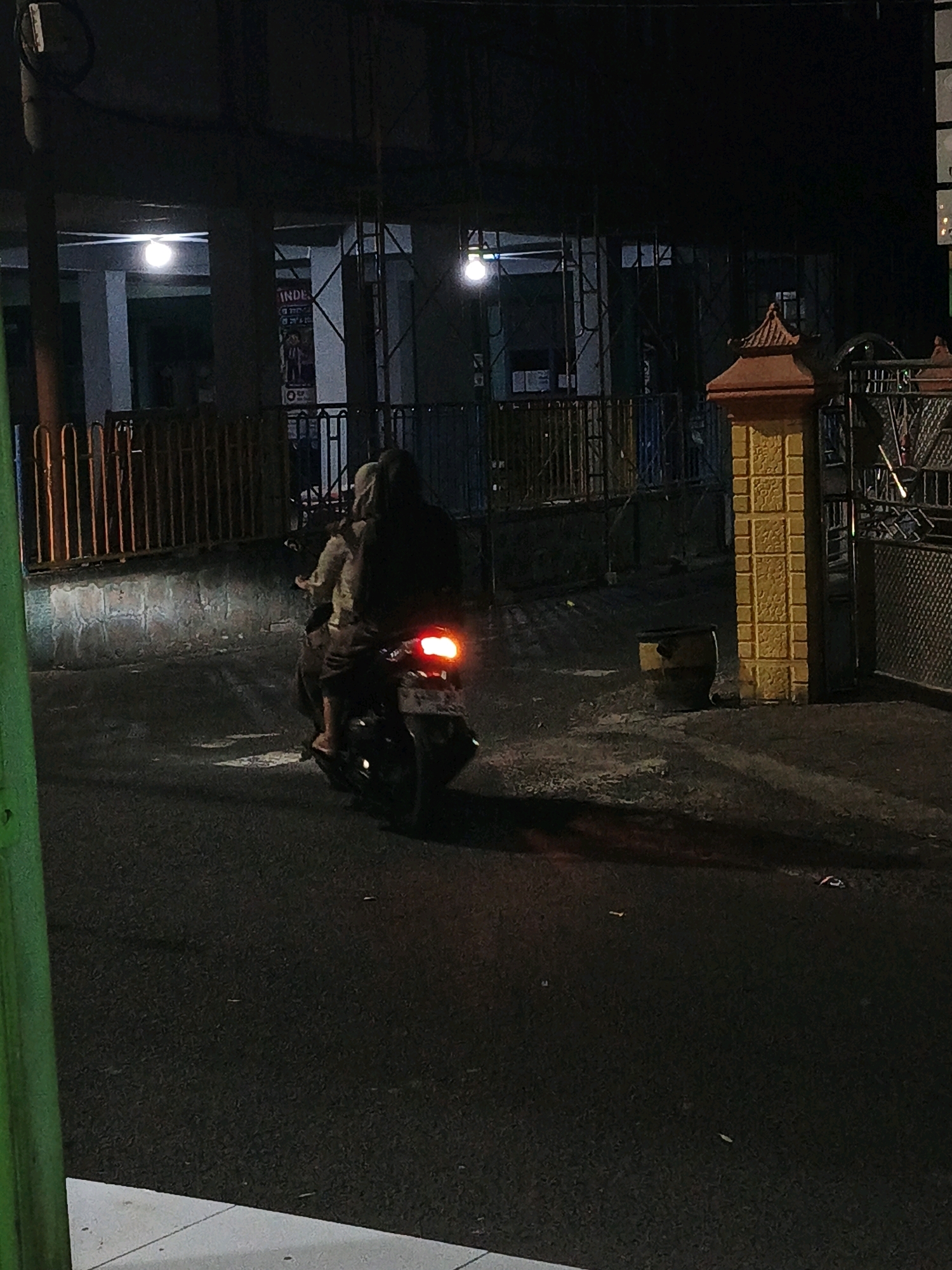 At night, on an asphalt road in Ketegan, Indonesia, two individuals are riding a motorcycle. The motorcycle's rear brake light is illuminated red. The individuals are wearing dark clothing. The rider is wearing a hood, and the passenger is wearing a light-colored shirt. The road surface shows some texture. To the left of the road, there is a metal fence with vertical bars, and behind it, a brick wall. Further back, there is a building with visible lights and some posters on a wall. To the right of the road, there is a decorative yellow pillar with a reddish-brown cap. Behind the pillar is a metal fence. The overall scene is dimly lit by artificial light sources.