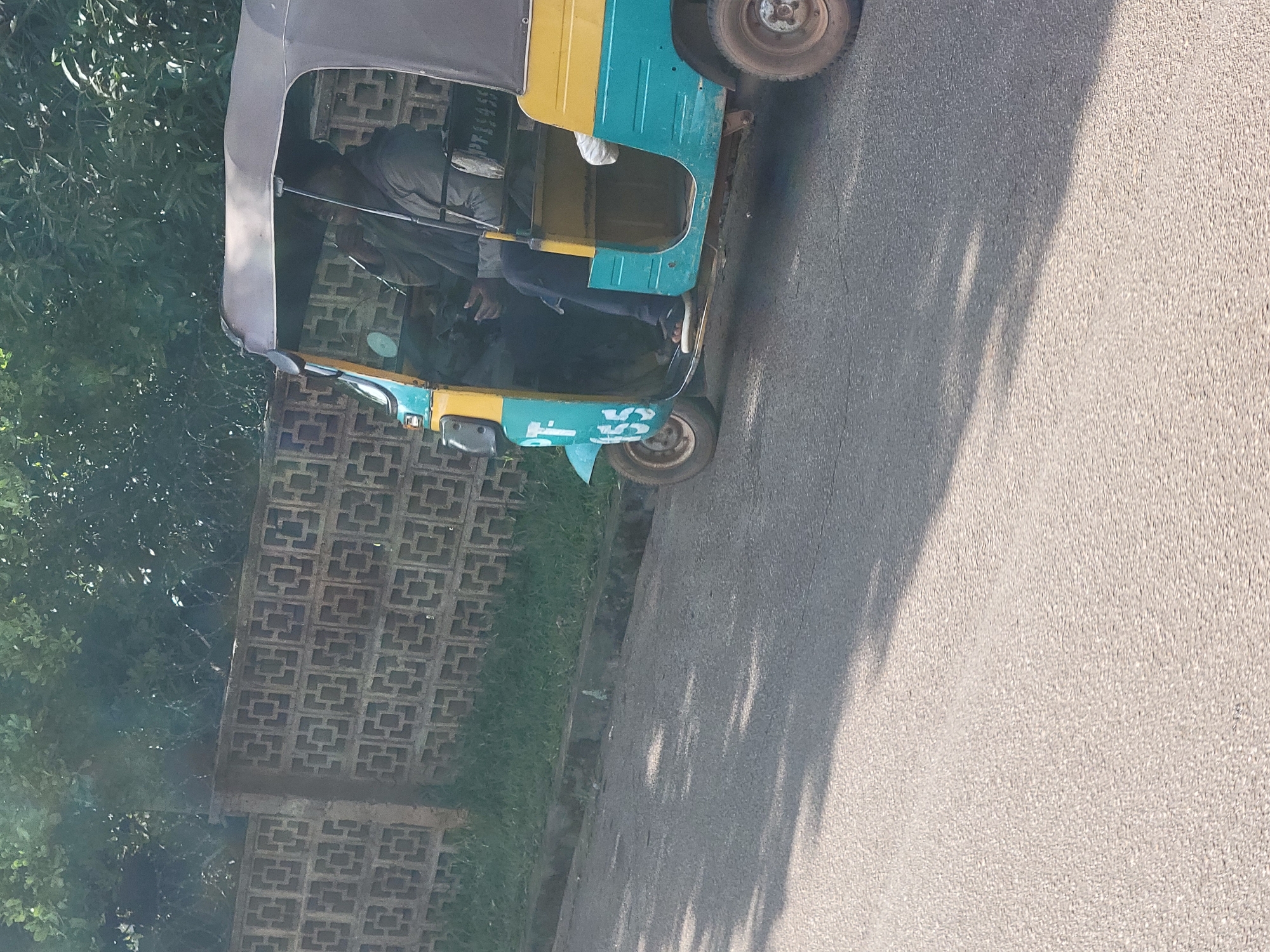 An auto rickshaw, painted teal and yellow, is parked on a paved road in Jos, Nigeria. The rickshaw has a grey canopy. Two individuals are visible inside the rickshaw. One person, likely the driver, is wearing a grey jacket and dark pants, and appears to be holding a phone. Another person, partially obscured, is seated behind the driver. The side of the rickshaw displays the text "PT" and "155" in white. In the background, a decorative concrete block wall runs along the side of the road, with lush green foliage behind it. The road surface is asphalt, with dappled sunlight casting shadows.