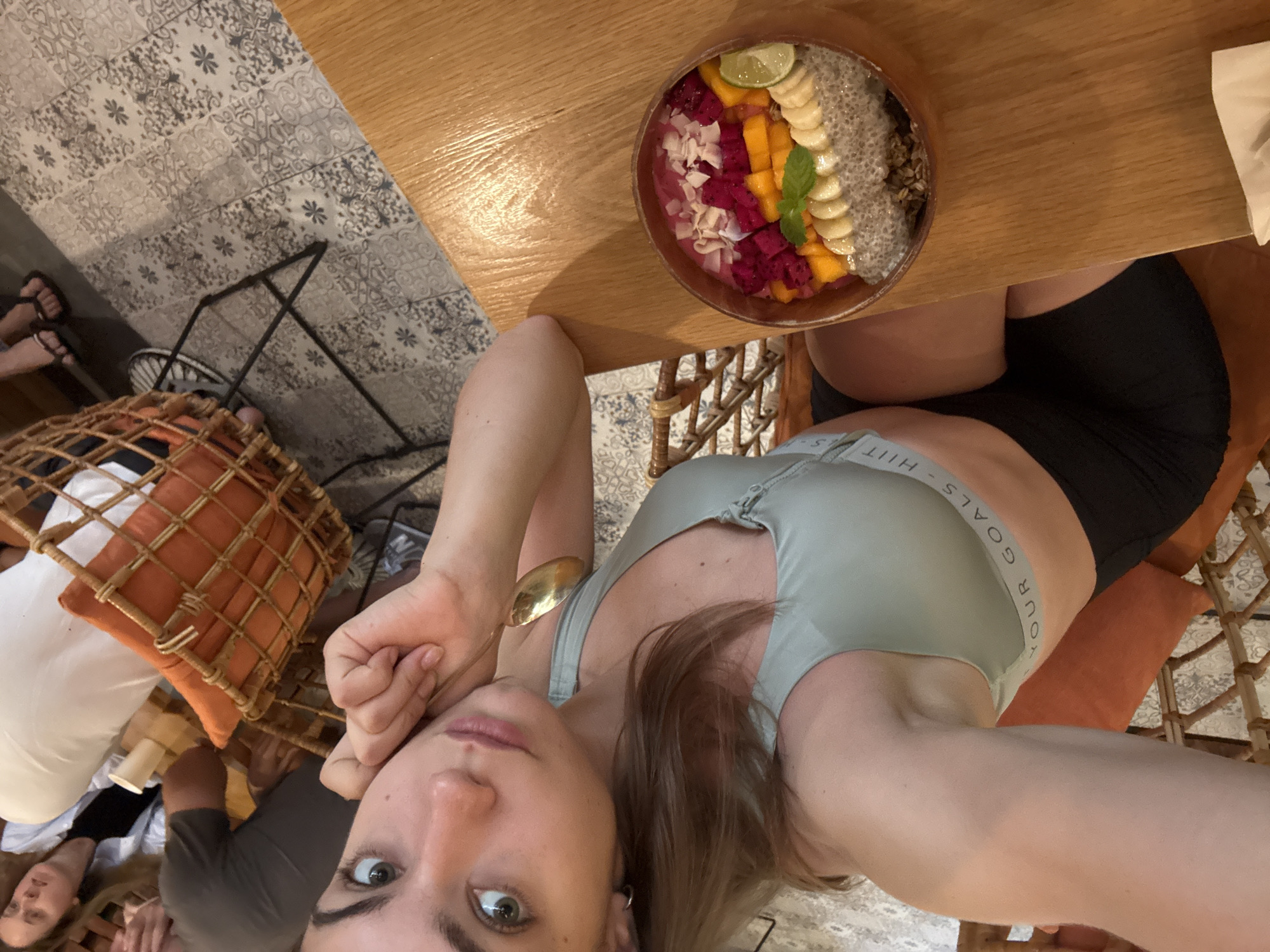 A selfie shot at an angle looking down from a seated person, most likely female. The person is fair-skinned and has light brown hair. The person wears a light green sports bra with "YOUR GOALS - HIIT" printed on the left side. The person is seated at a light wood table. On the table is a bowl containing dragonfruit, mango, banana, chia seeds, and coconut flakes. The person is holding a gold-colored spoon. There are other people seated to the left of the person. The seated area appears to be indoors. The floor is patterned with grey tiles. There are woven wicker chairs and orange-colored cushions. This location is likely in Đà Nẵng, Vietnam.