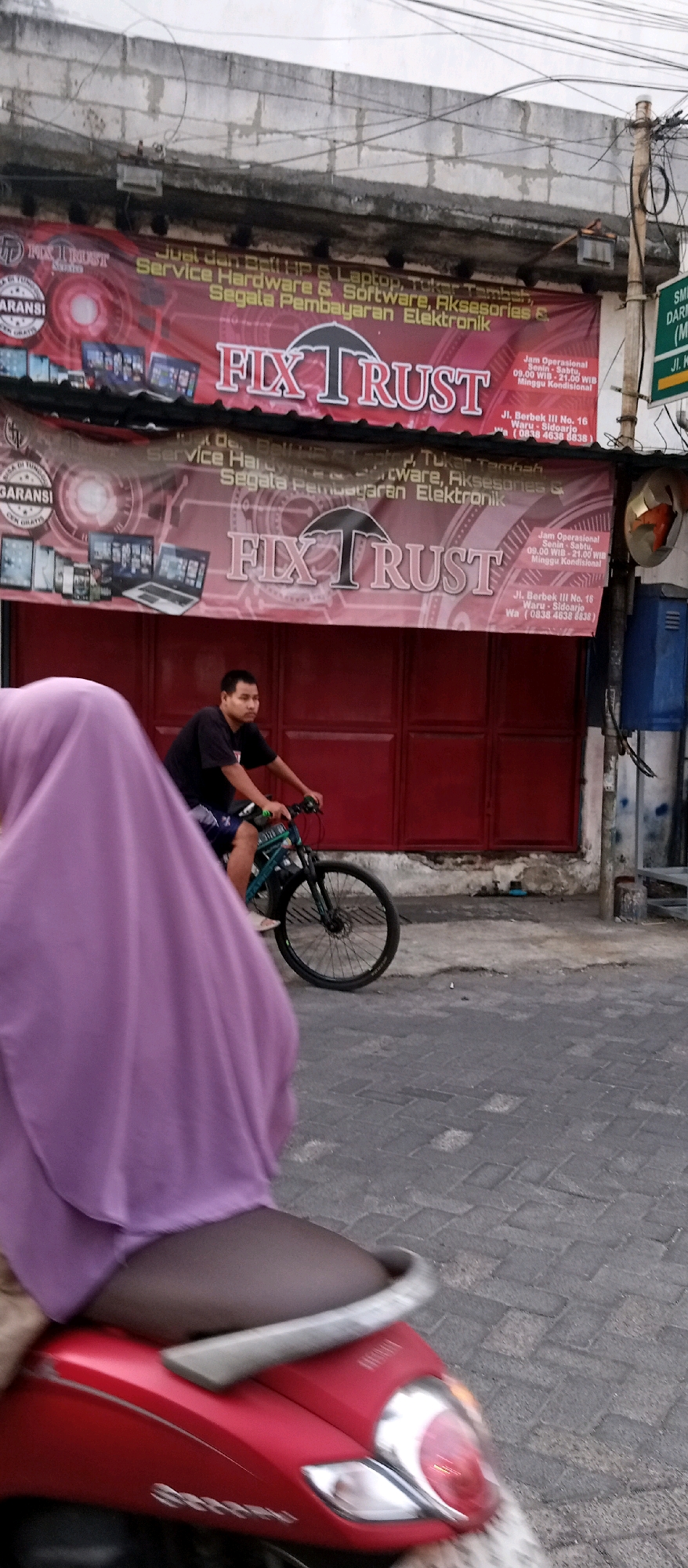 A man rides a bicycle in front of a storefront in Wadungasri, Indonesia. The man is wearing a black t-shirt and blue shorts and is looking forward as he pedals. The storefront is decorated with a large banner that reads "FIX TRUST" and advertises services such as computer repair and accessories. A red scooter is parked in the foreground, with its seat and rear end visible. The street is paved with cobblestones. The scene appears to be during the daytime, with natural light illuminating the area.