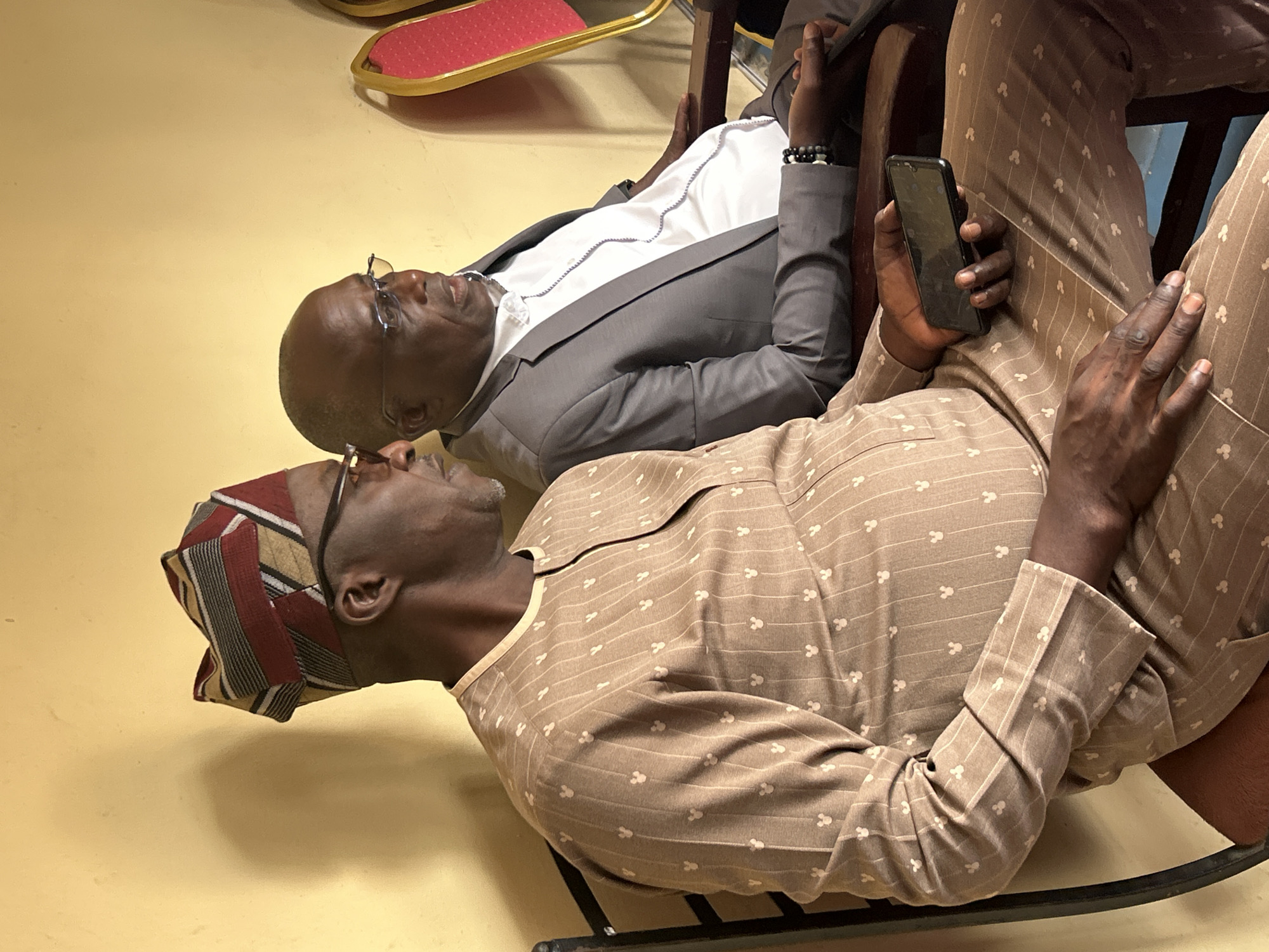 Two dark-skinned men are seated in chairs in an indoor setting with a light yellow floor. The man in the foreground is wearing a brown and white patterned shirt and matching pants, along with a brown and maroon cap. He is holding a black smartphone. The man behind him is wearing glasses and a grey suit over a white shirt. They are both turned to look in the same direction. Part of a chair with a red seat is visible in the background. The location is likely in the city of Jos, Nigeria.