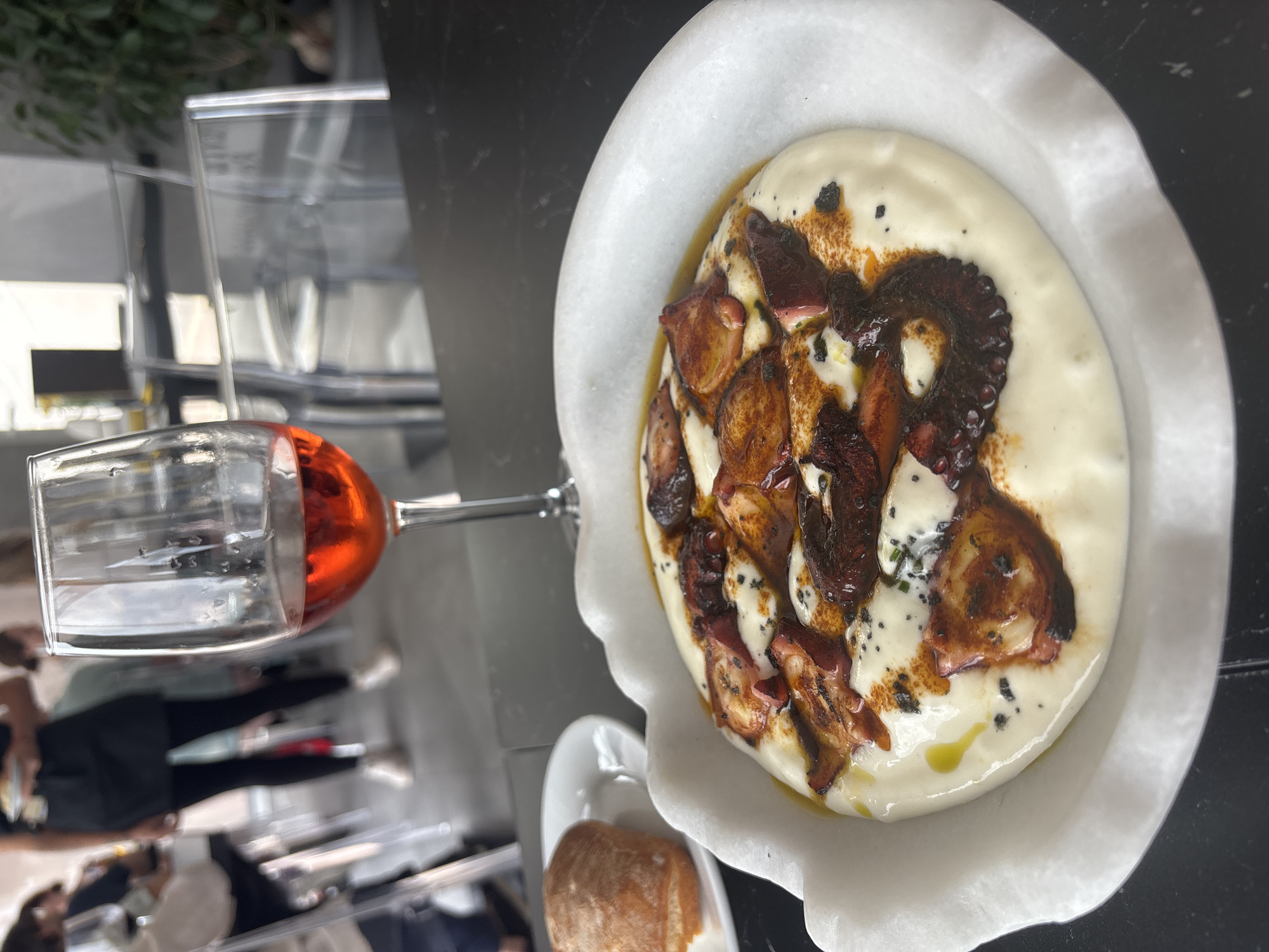 A close-up, eye-level shot shows a dish of octopus served in a white, scalloped bowl with a creamy white sauce and garnished with dark spices and olive oil. To the left of the bowl, a white plate holds a small, round bread roll. In the background, a glass of rosé wine is positioned behind the food. Further back, blurred figures of people are visible, and transparent chairs suggest an indoor dining setting, possibly a restaurant in El Parador de las Hortichuelas, Spain. The lighting is bright, casting a natural glow on the food and wine.