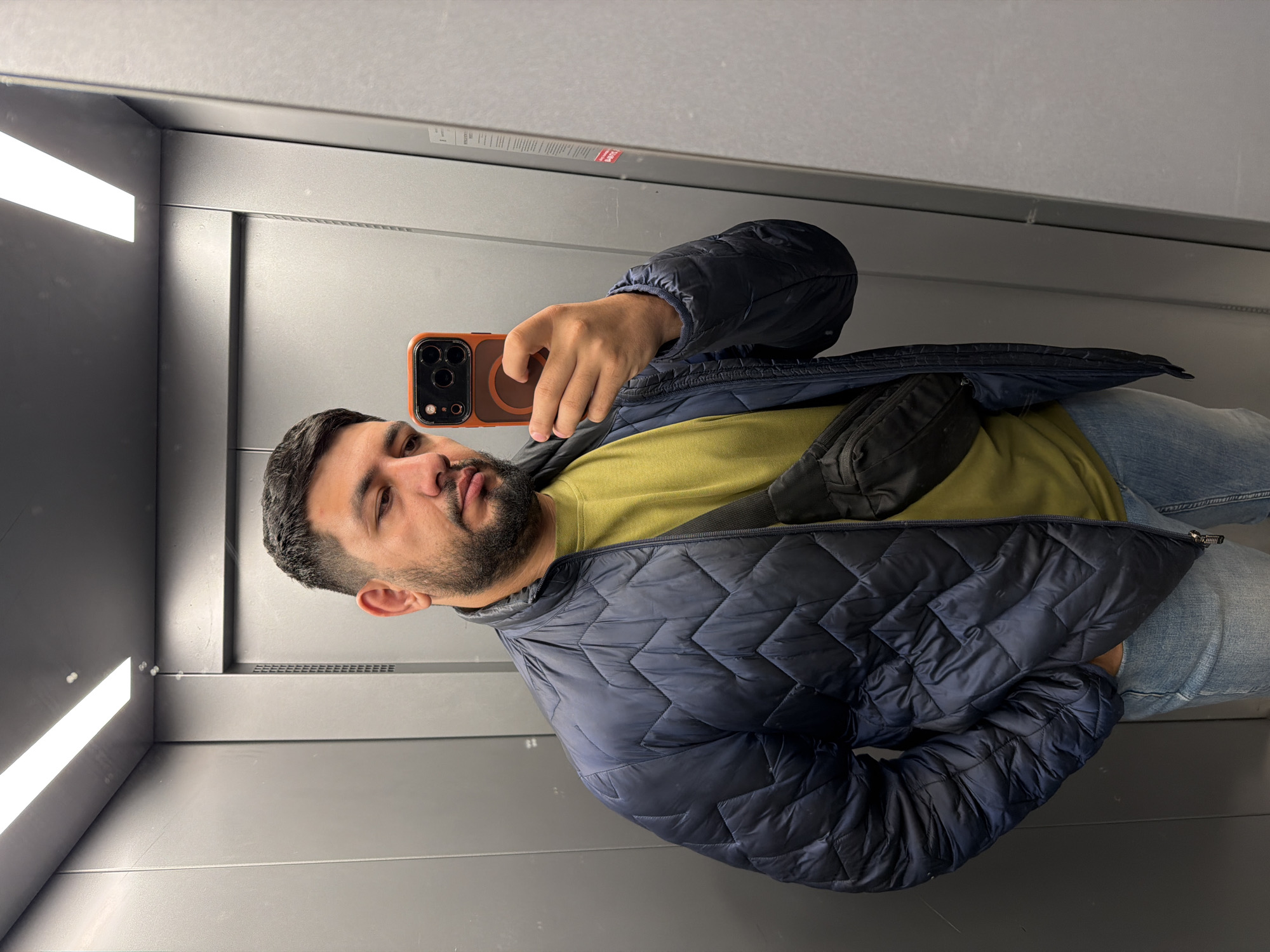 A man is taking a selfie in what appears to be an elevator. He has dark hair and a beard, and he is wearing a yellow shirt, blue jeans, and a navy blue puffer jacket. He is holding a smartphone with an orange case in his right hand. The elevator has gray walls and a bright fluorescent light. The location is assumed to be Krasnoyarsk, Russia.