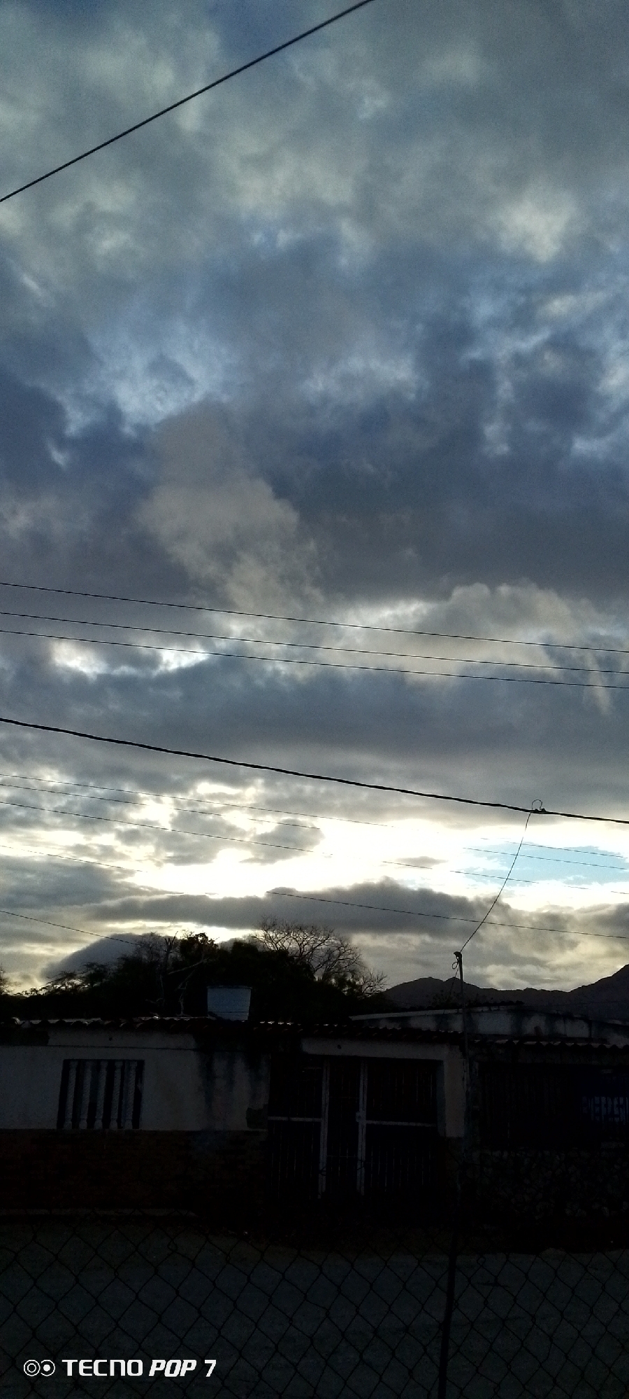 The image shows a low-angle view of a cloudy sky over a partially obscured building and landscape, likely taken in Altagracia, Venezuela. Overcast, dark clouds dominate the sky, with brighter areas peeking through, suggesting a possible sunset or sunrise. Several electrical or telephone wires stretch across the sky.
In the foreground, a blurred wire fence runs horizontally. Behind the fence is a one-story building, partially hidden by shrubbery. The building has barred windows and a door, and its facade appears weathered. The lighting is dim, making it difficult to discern details, but a mountain range is faintly visible in the background. The "TECNO POP 7" watermark is in the lower-left corner.