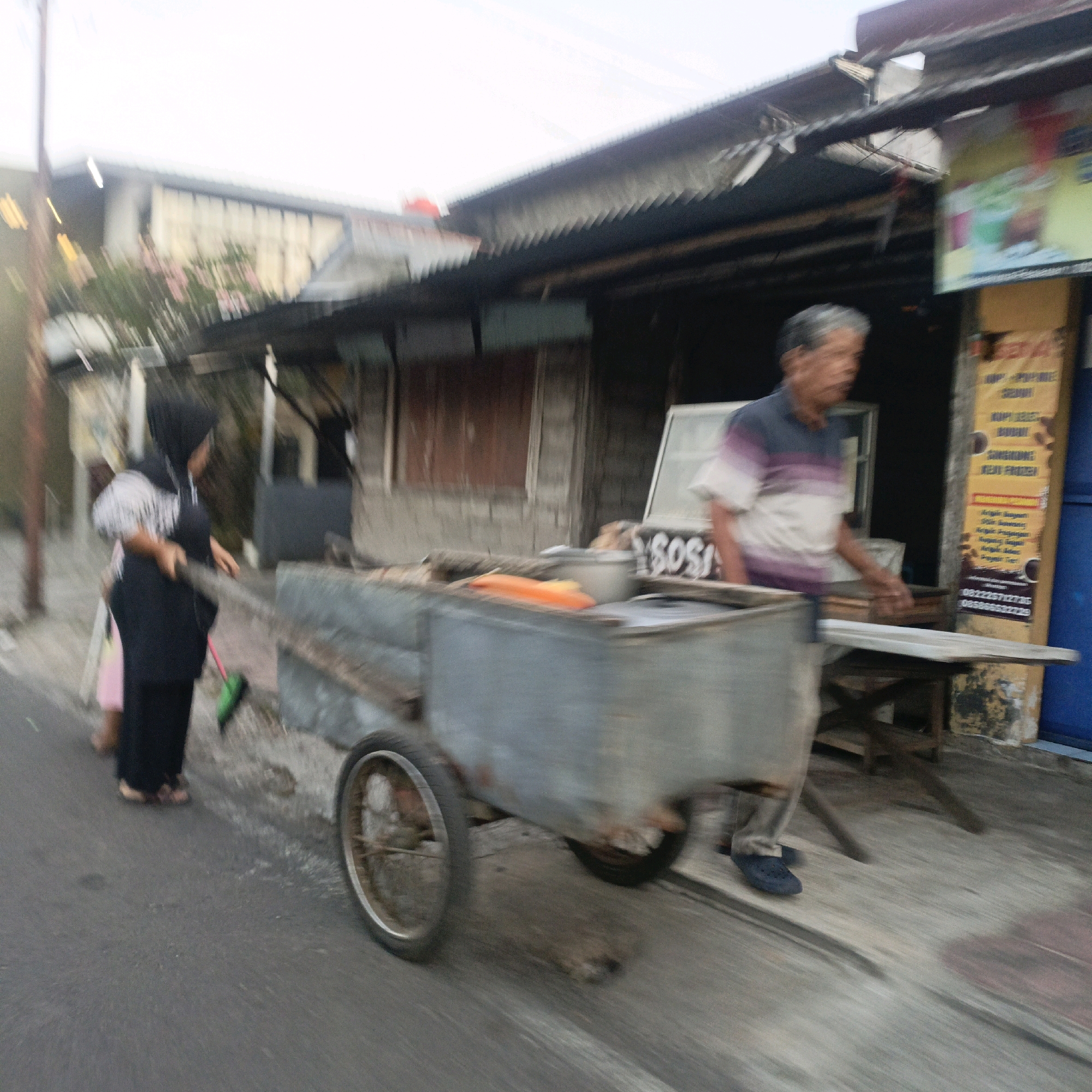 A motion-blurred, medium-distance, eye-level, outdoor shot captures a street scene in Salatiga, Indonesia. In the foreground, a person wearing a black hijab and dark clothing pushes a metal cart with a green broom leaning against it. The cart contains a pot and an orange item, possibly food. Behind the cart, a person in a horizontally striped shirt, appearing to be an older male, stands near a cooler and a wooden structure. A yellow sign with Indonesian text is visible on the right side of the frame. The background depicts a weathered building with a corrugated metal roof. The ground is asphalt and concrete, and the overall scene suggests a commercial or residential street.