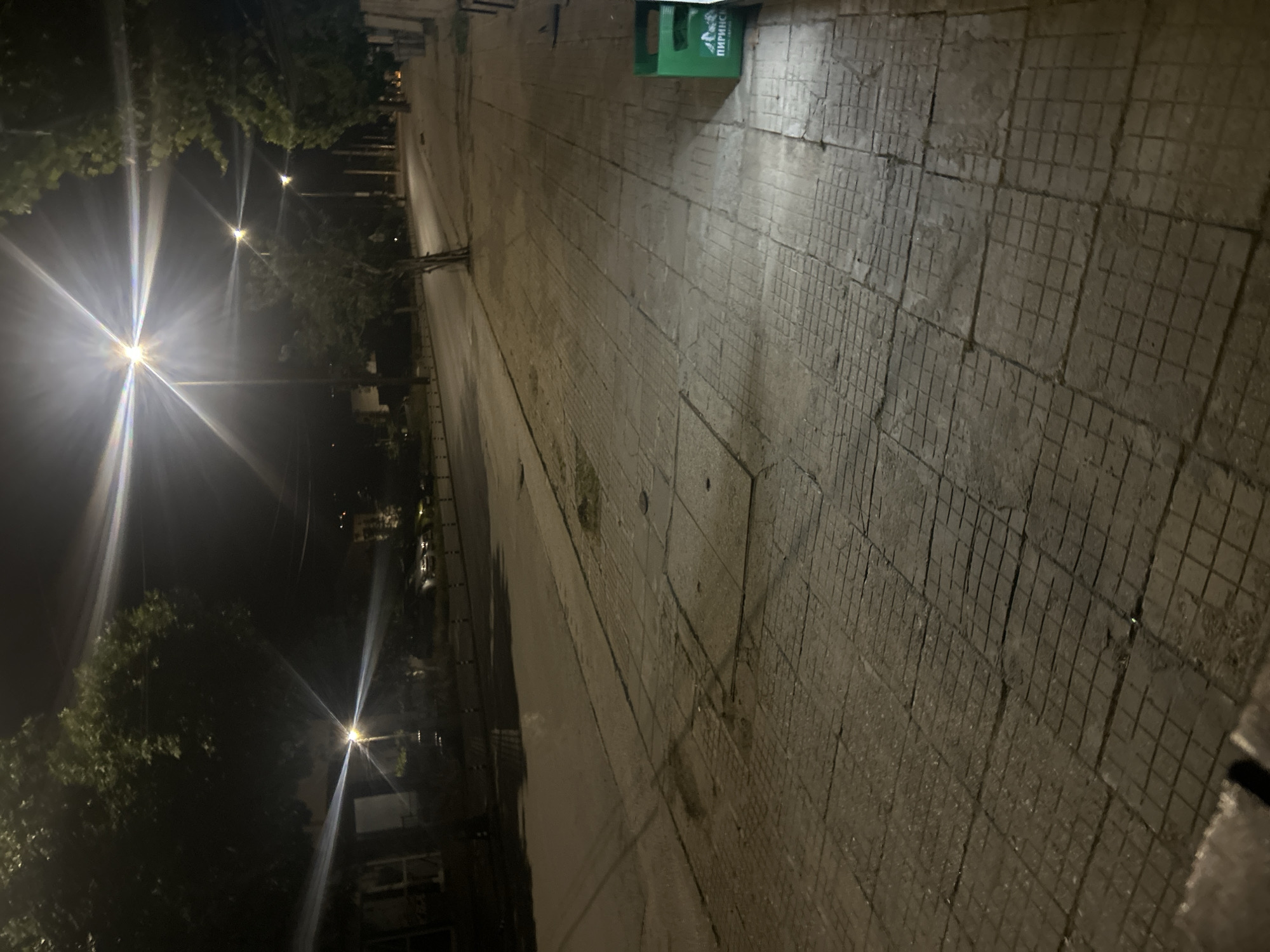 The image depicts a quiet urban street scene at night in the city of Unknown, Unknown. The perspective is from a low angle on a sidewalk, looking down a street illuminated by multiple streetlights.
The scene is dominated by artificial lighting, with at least four streetlights clearly visible, each emitting a bright, starburst-like flare. These lights cast a warm yellow-white illumination across the asphalt street and the concrete-tiled sidewalk. The sky is not visible due to the presence of trees and the framing.
In the foreground and extending to the right, a worn concrete sidewalk is visible, constructed from various rectangular tiles. Many of these tiles feature a grid-like pattern, while others are smoother and appear to be utility covers embedded in the pavement. Patches of what appears to be dirt or sparse vegetation are visible in cracks and along the edges of the sidewalk.
To the left of the sidewalk, an asphalt road stretches into the distance, gently curving to the right. A line of short, dark bollards or posts borders the road. On the far side of the road, several parked cars are discernible, though their details are indistinct in the low light.
Trees with green foliage line both sides of the street, obscuring distant buildings and parts of the background. A slender, young tree trunk grows directly from a gap in the sidewalk near the center of the image.
On the far right, in the mid-ground, a bright green plastic crate stands upright against a dark vertical surface, possibly a building wall. The crate has text visible on its side in Cyrillic script, which reads "ПИРИНСКИ".
No individuals, animals, or active human interactions are present in the scene. The overall impression is one of stillness and the artificial glow of an urban night.