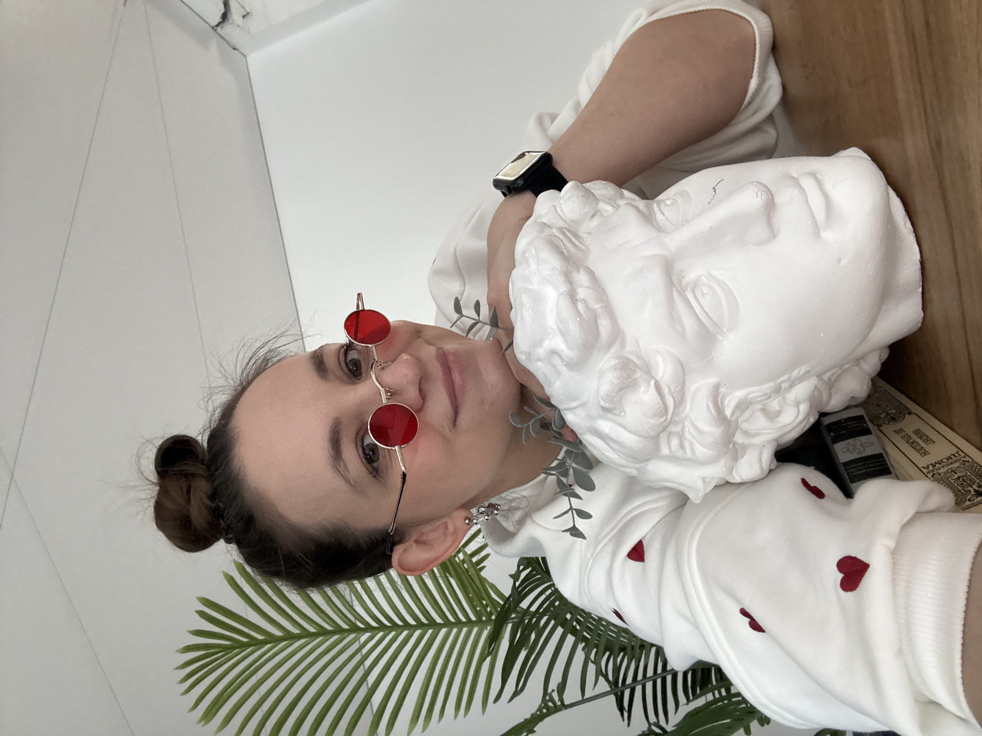 A person with brown hair tied in a bun and wearing red-tinted round glasses is holding a white plaster bust of a human head against their shoulder. The person is wearing a white hooded sweatshirt adorned with small red hearts. The person has a black smartwatch on their left wrist. A leafy green plant is visible near the bottom of the frame. The background includes a white wall and a wooden surface. The image was taken indoors in a space located in the city of Chaikovsky, Russia.